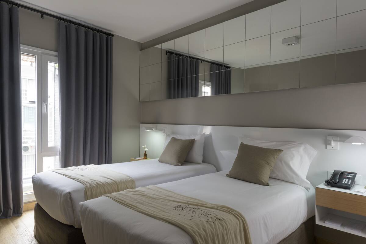 Une chambre d’hôtel avec deux lits, un grand miroir, une table de chevet avec un téléphone et une fenêtre avec des rideaux.