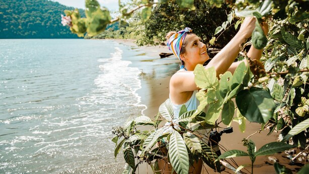 A person standing by a beach with a view of the sea and a forested hill in the background.