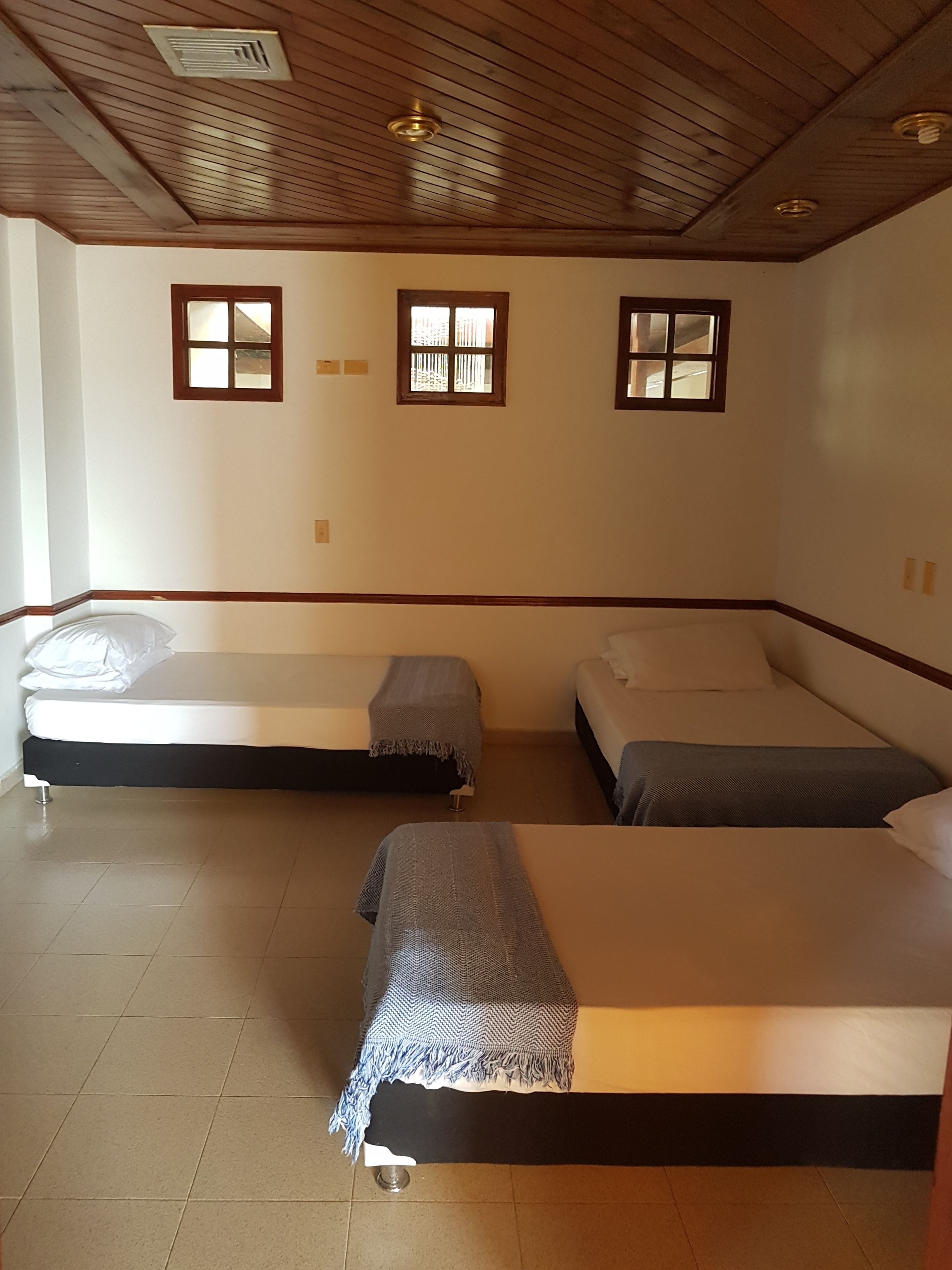 A room with two beds, wooden ceiling, and three windows.
