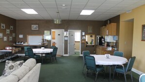 A room with a green carpet, white tables, and teal chairs. There is a whiteboard, a refrigerator, and a microwave on the wall.
