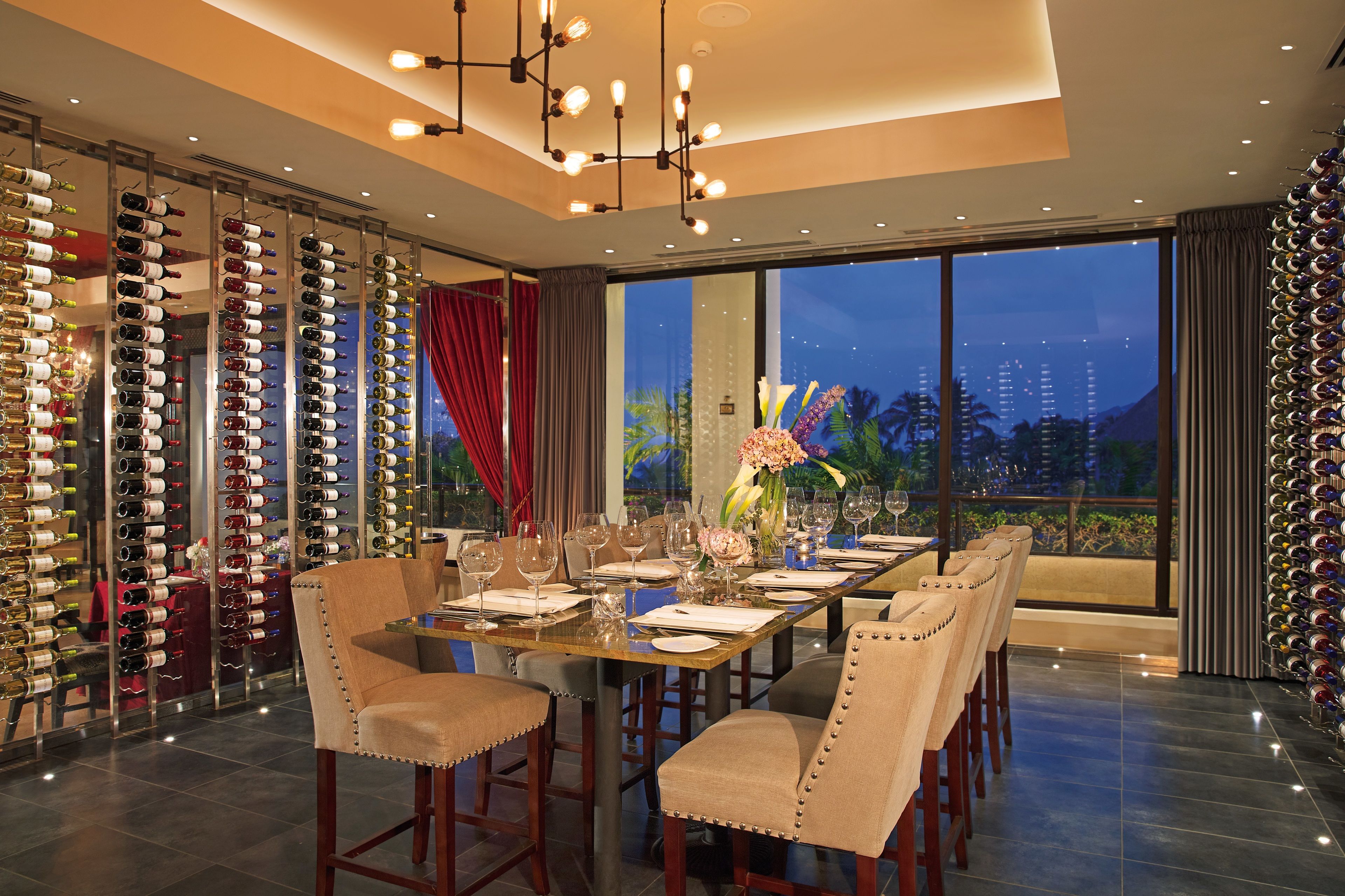 A dining area with a long table set for six, surrounded by wine racks, and a view of the city at dusk.