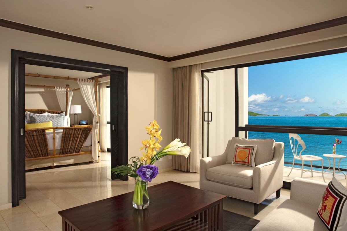 A hotel room with a bed, a sitting area with a chair and a table, a vase with flowers, and a view of the sea.