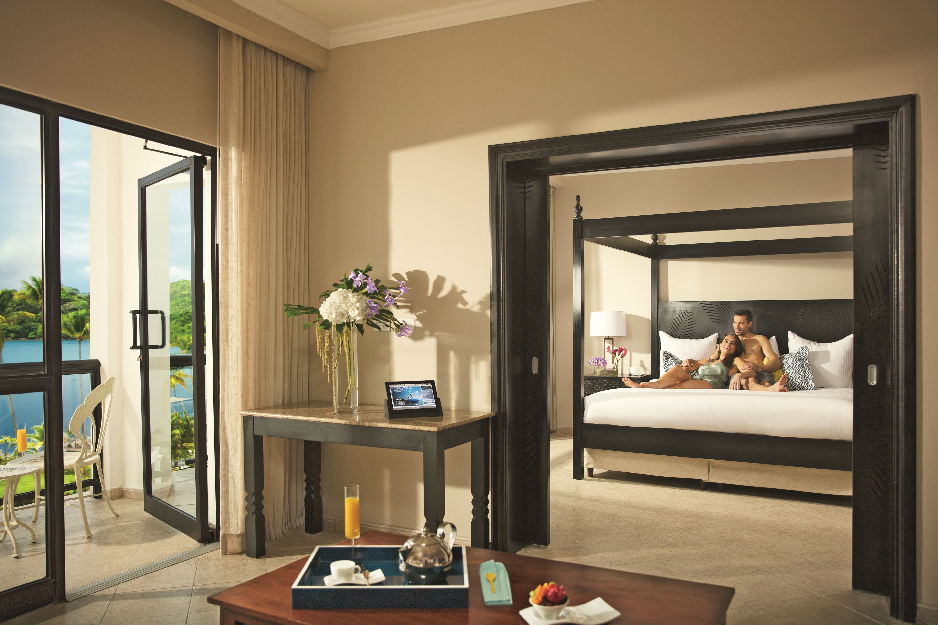 A hotel room with a bed, a table with a tray of breakfast items, a laptop, and a view of the outdoors.