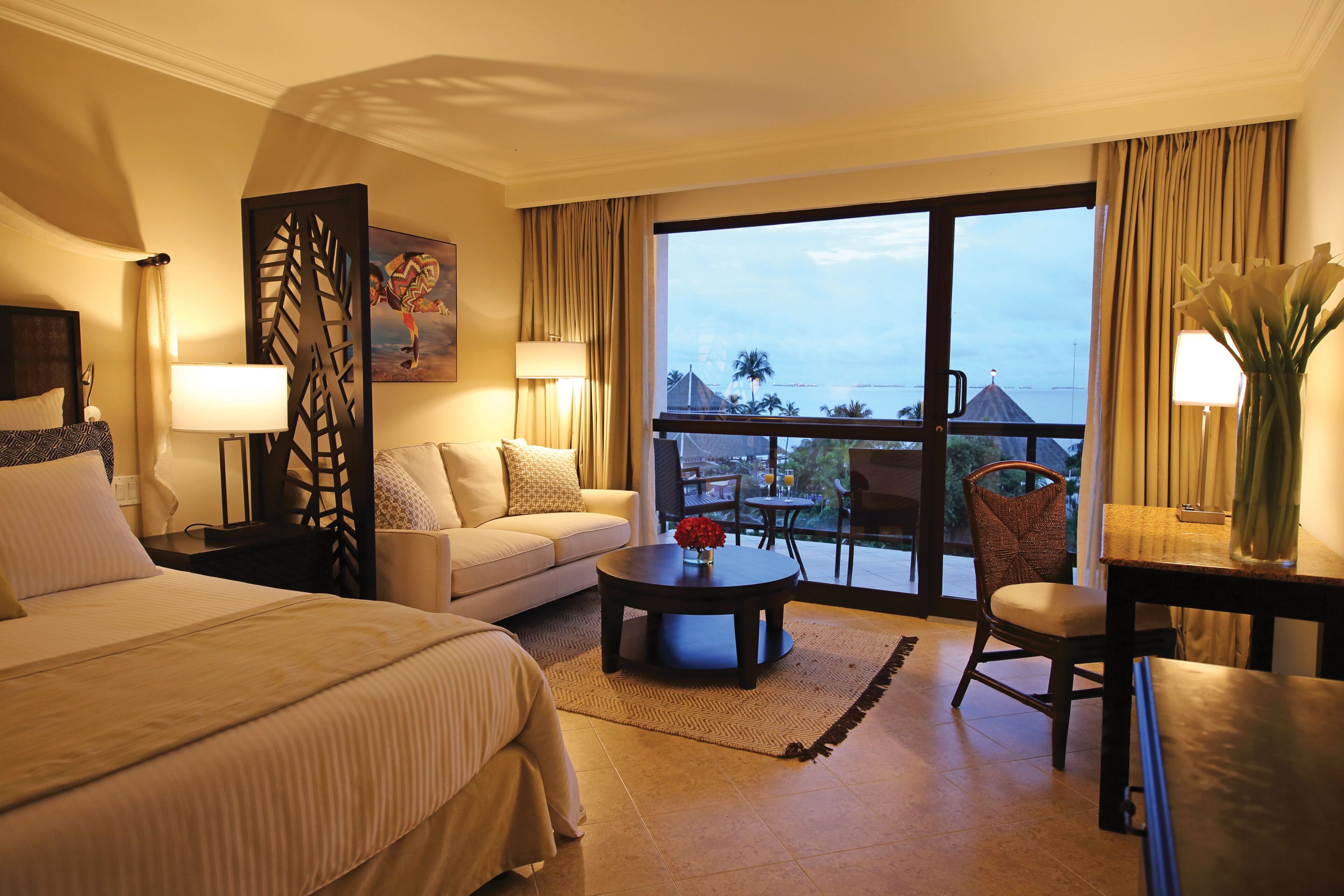 Une chambre d’hôtel comprenant un lit, un canapé, une table basse, une chaise, un bureau, une lampe, un tableau et une vue sur l’extérieur.
