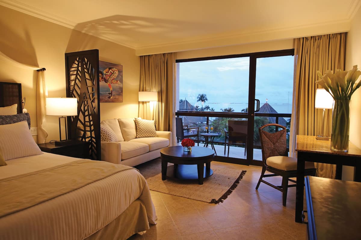 A hotel room with a bed, a sofa, a coffee table, a chair, a desk, a lamp, a painting, and a view of the outdoors.
