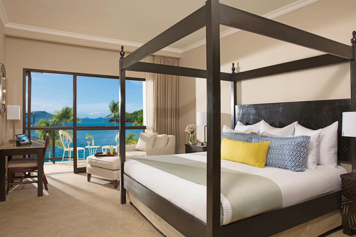 A four-poster bed with a view of the ocean, a desk with a laptop, and a chair by the window.