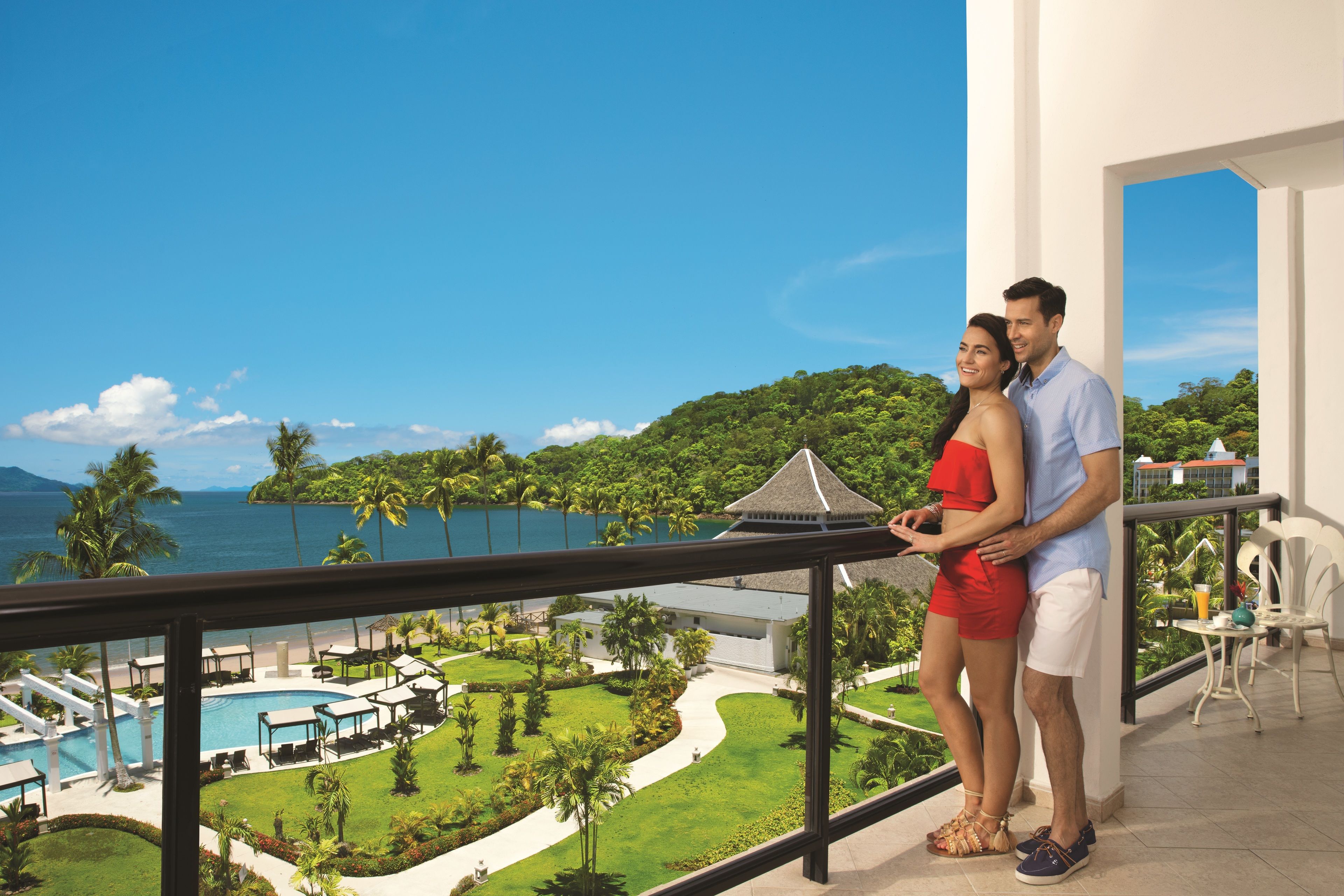 A couple standing on a balcony overlooking a pool and lush greenery with a view of the sea.