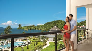 A couple standing on a balcony overlooking a pool and lush greenery with a view of the sea.