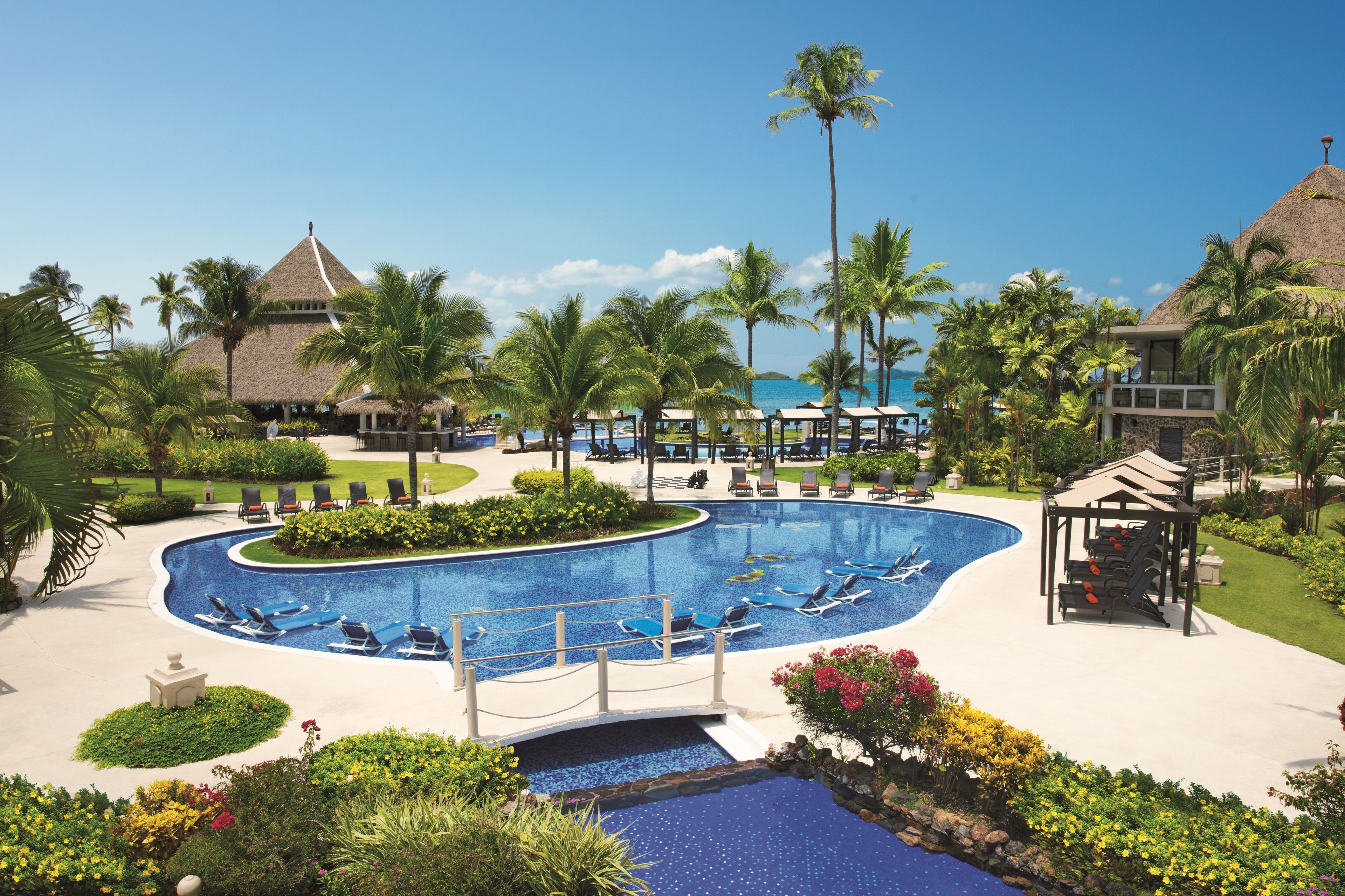Un resort con una piscina grande, sillones de descanso, palmeras y un edificio de techo de paja.