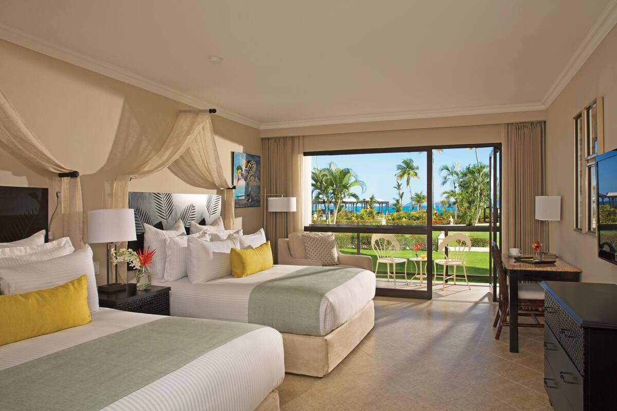 A hotel room with two beds, a balcony with a view of palm trees, and a dining area.