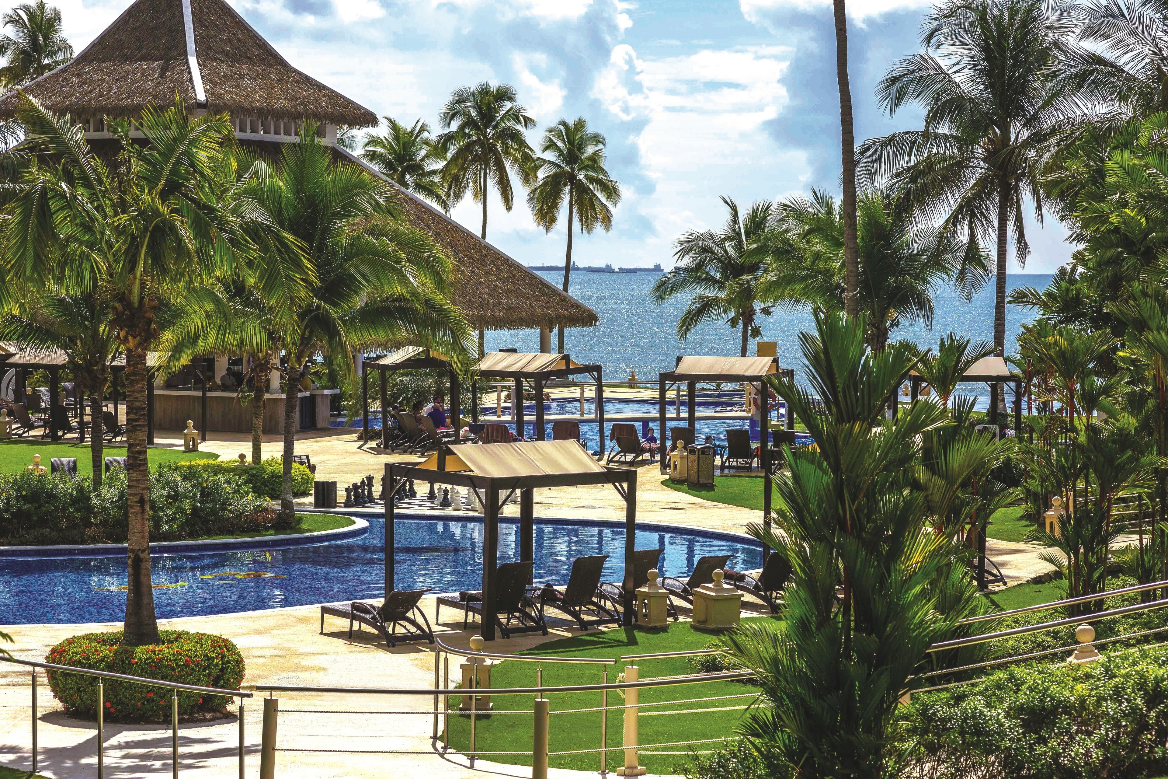 A resort with a pool, lounge chairs, palm trees, and a view of the sea.