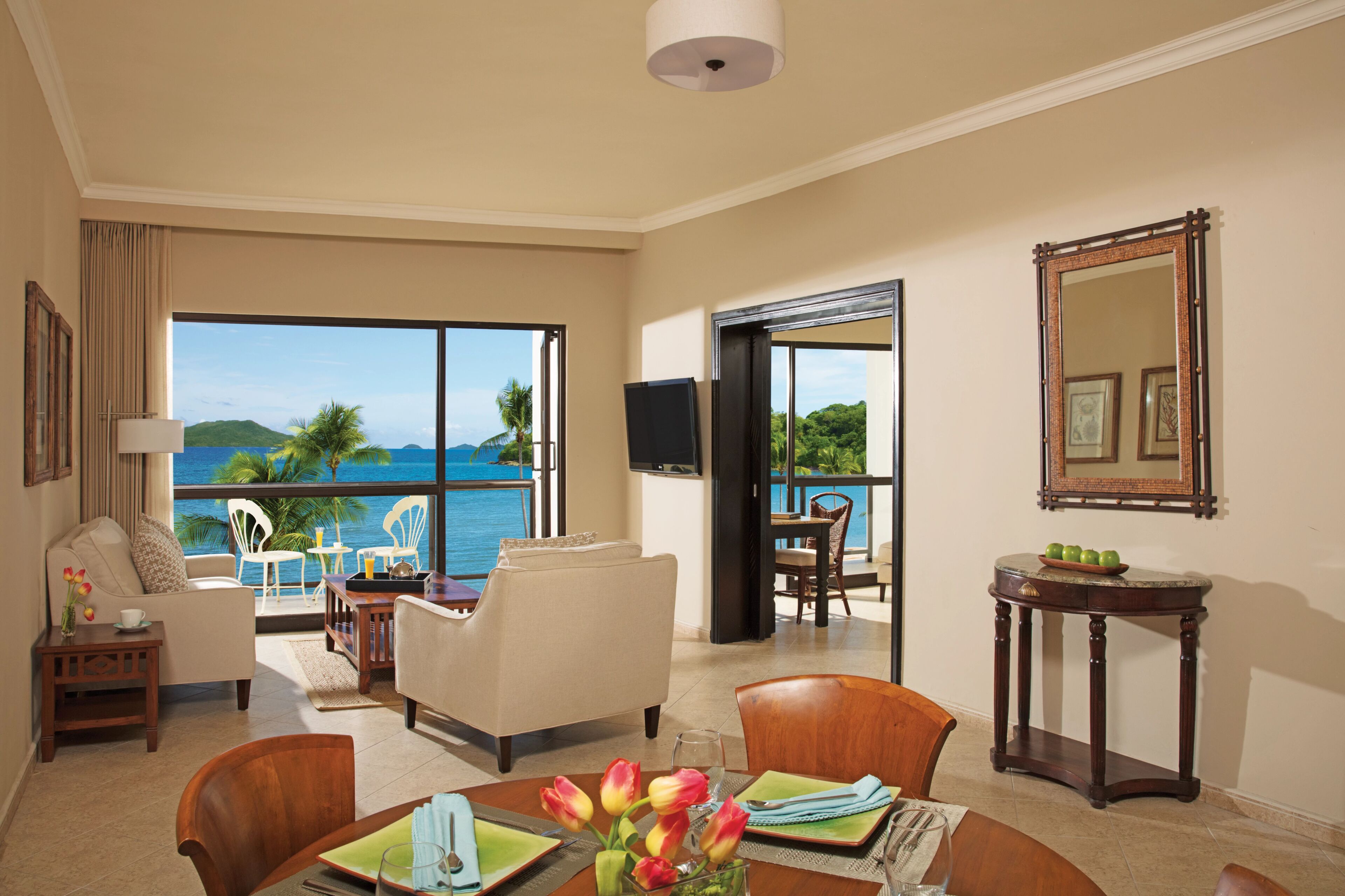 A spacious room with a balcony overlooking a scenic ocean view, a dining area with a table set for two, and a television mounted on the wall.