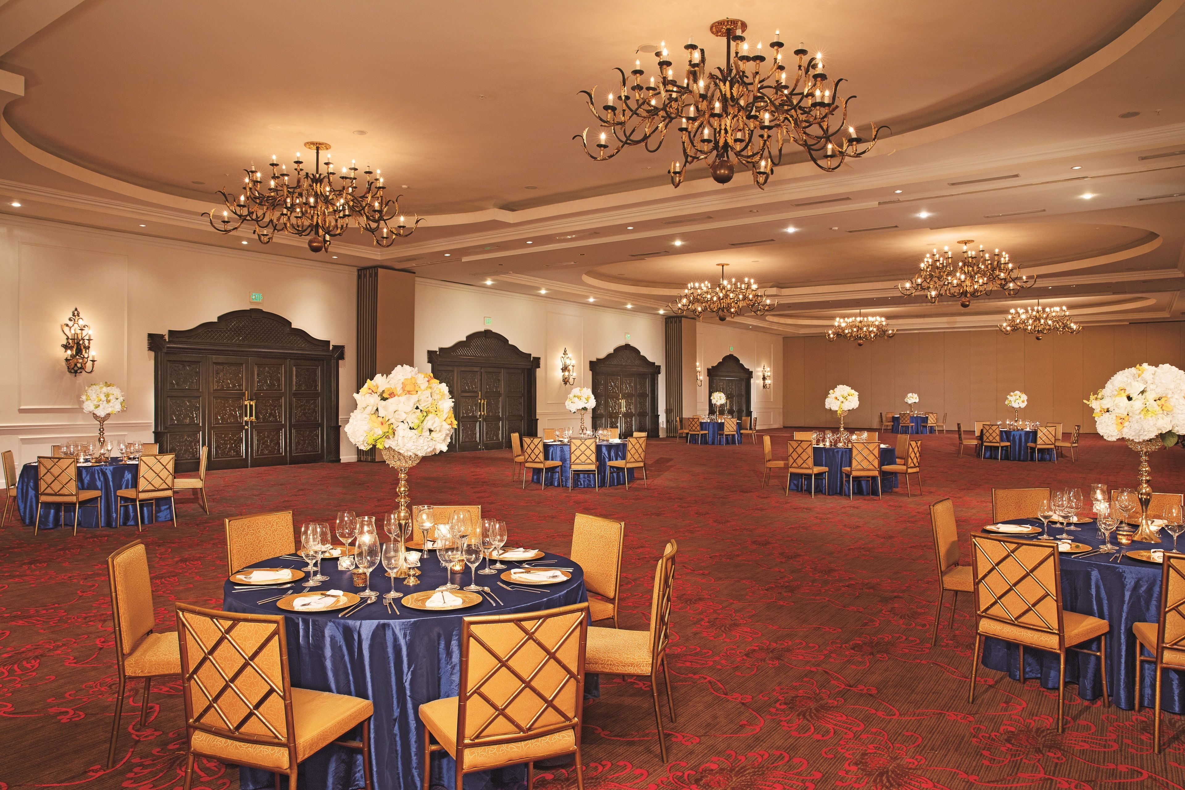 Un salón de banquetes con mesas redondas dispuestas para recibir comensales, con sillas doradas y manteles azules, candelabros y centros de mesa florales.