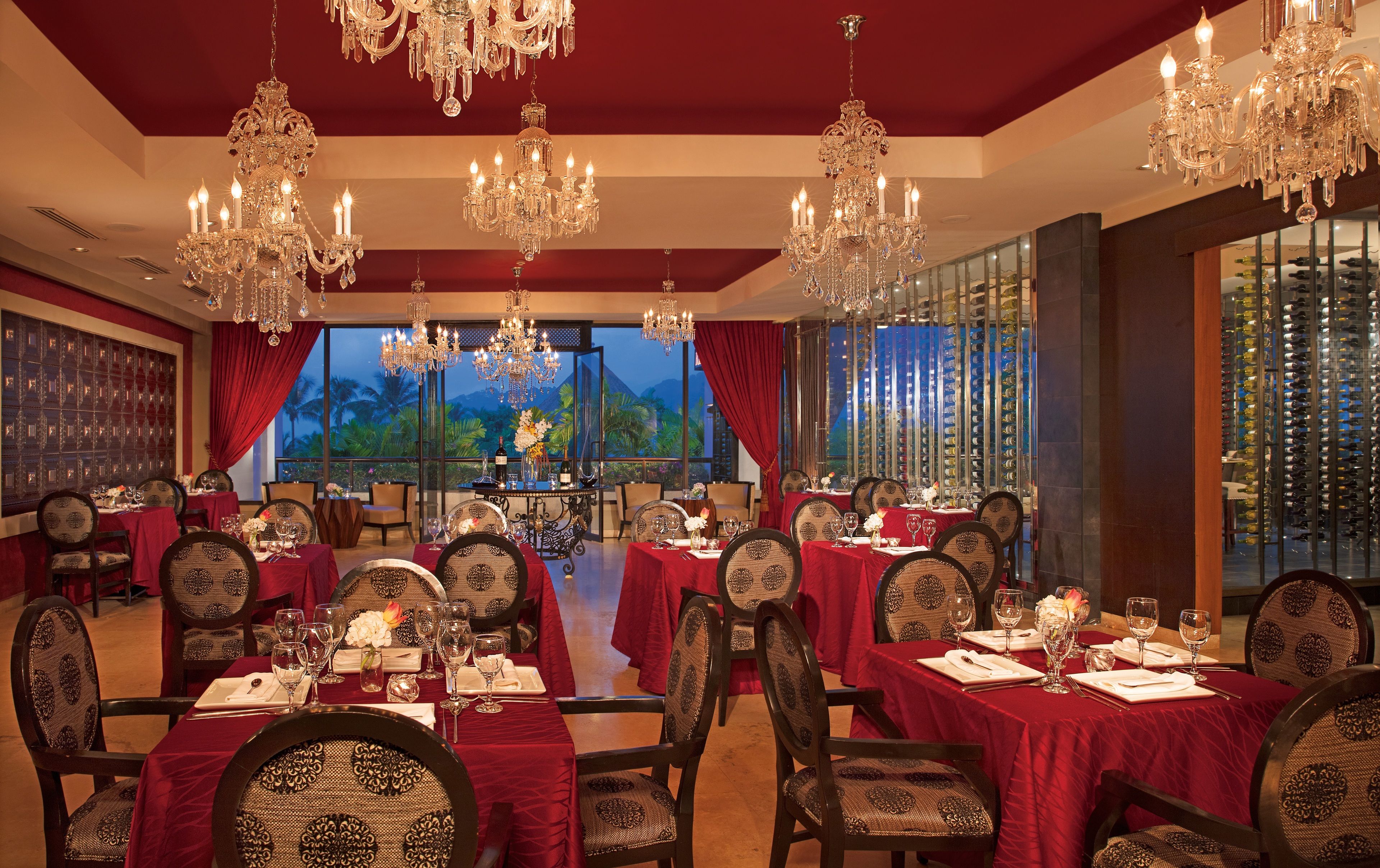Un comedor formal con varias mesas dispuestas para una comida, grandes candelabros y vistas a palmeras a través de las ventanas.