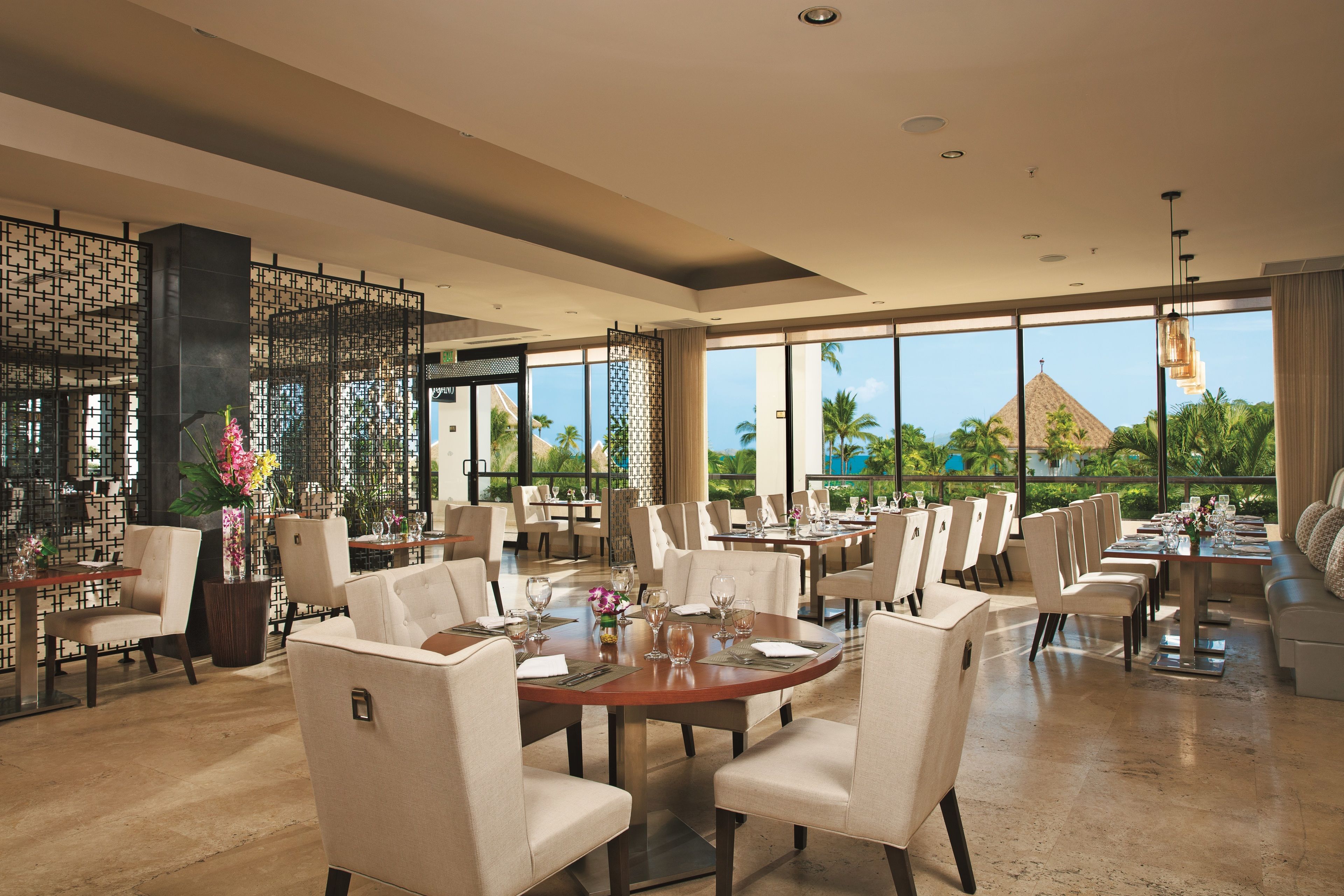 A spacious restaurant with high ceilings, large windows, and a view of palm trees and a pyramid. Tables are set with white chairs and centerpieces.