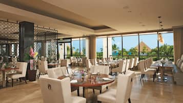 A spacious restaurant with high ceilings, large windows, and a view of palm trees and a pyramid. Tables are set with white chairs and centerpieces.