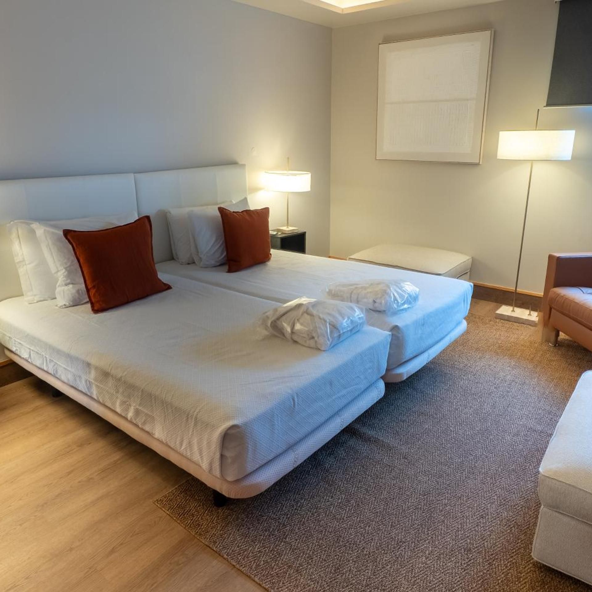 Habitación de hotel con cama doble, una mesita de noche con lámpara, otra mesita de noche más pequeña con lámpara, una silla y un ventanal grande.
