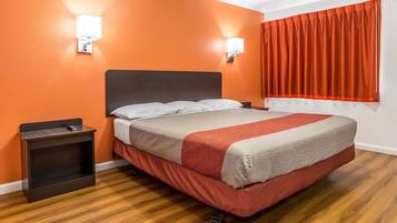 Ein Hotelzimmer mit einem Bett, zwei Wandleuchten, einem Fenster mit orangefarbenen Vorhängen und einem Holzboden.