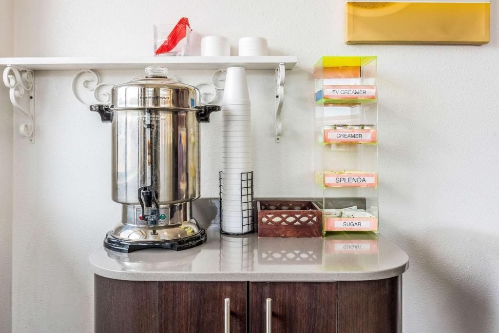 A kitchenette with a coffee pot, a white mug, and a shelf with labeled containers of creamer, sugar, and splenda.