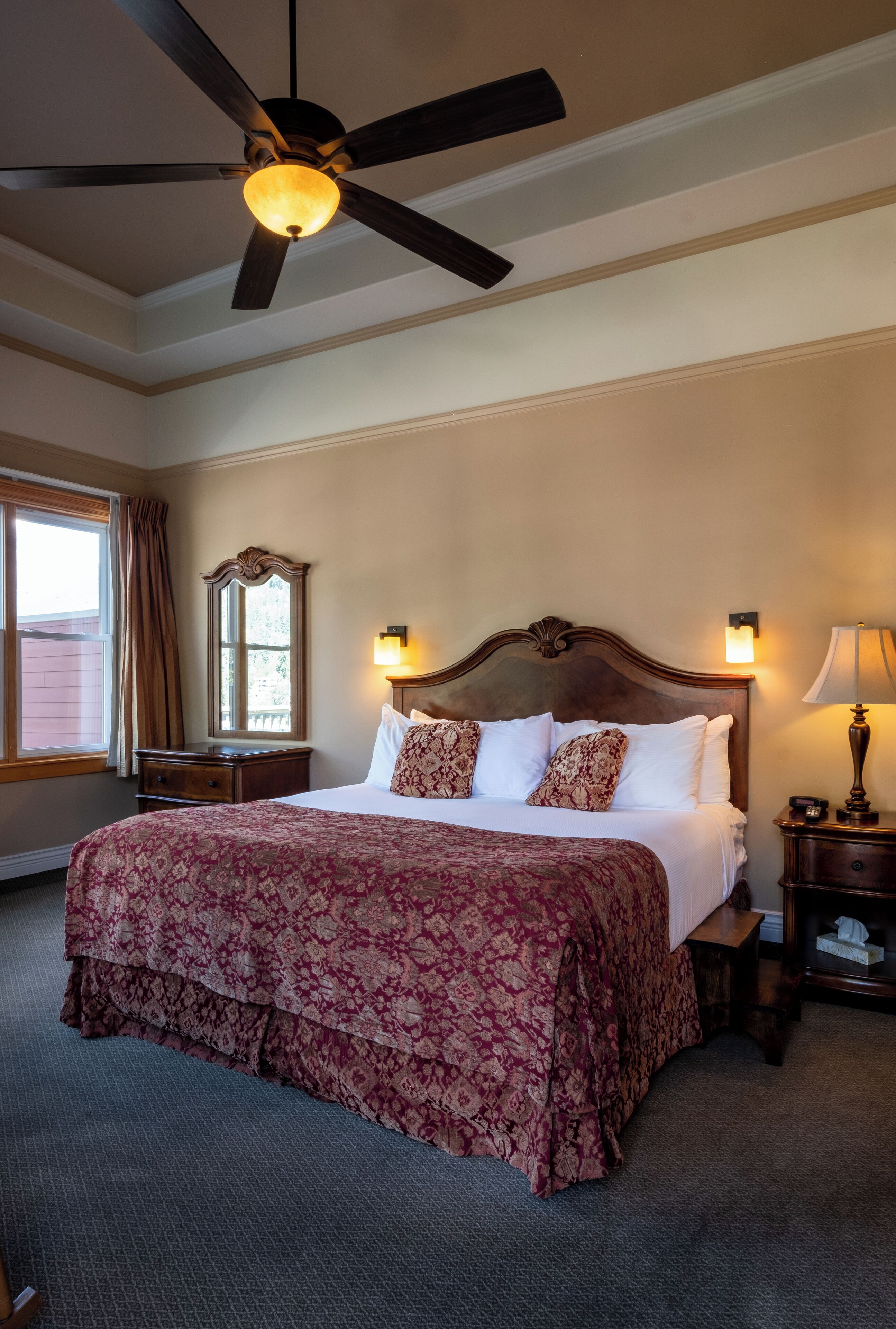 A hotel room with a large bed, a ceiling fan, a nightstand with a lamp, a window with curtains, and a mirror.