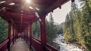 Un puente de madera con barandas rojas que lleva a un río rodeado de bosque denso.