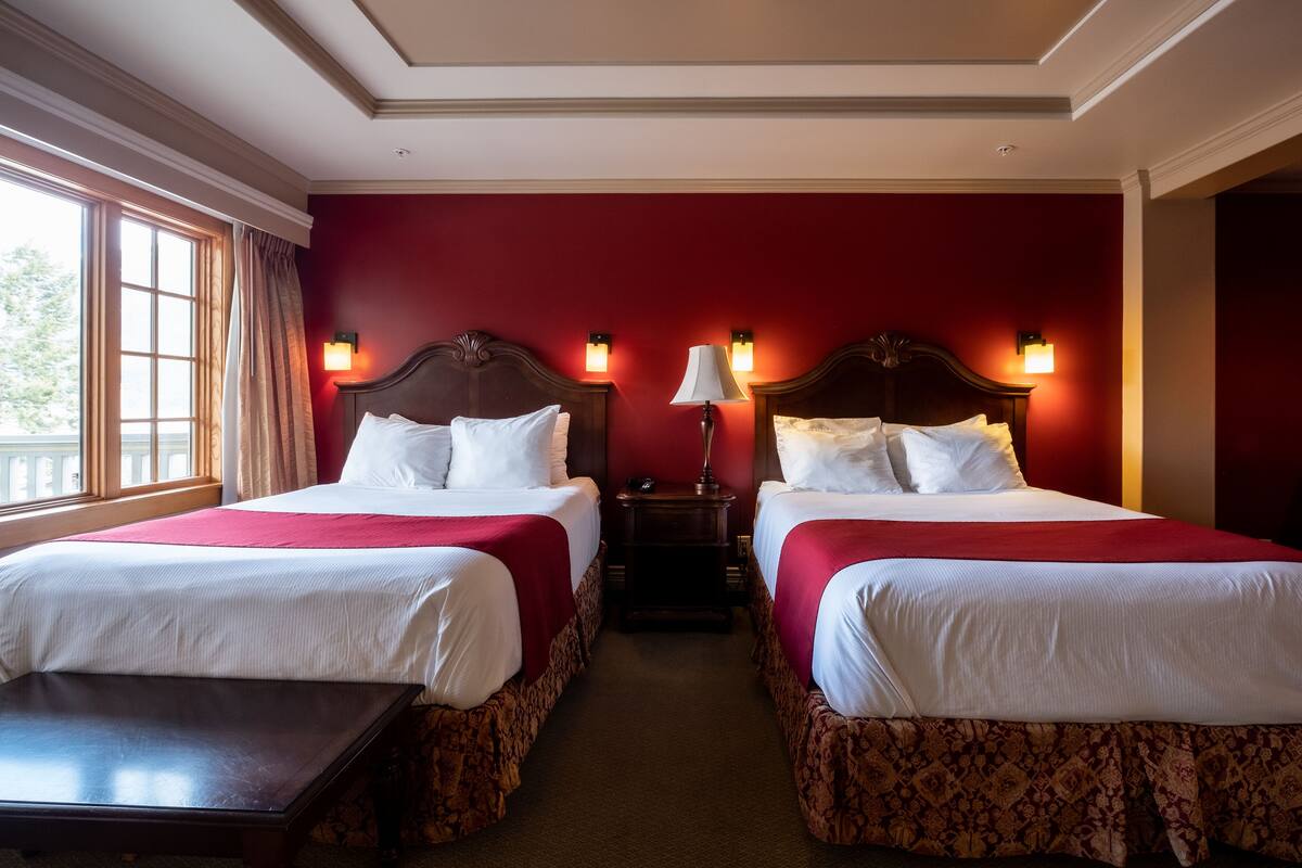 Habitación de hotel con dos camas, una mesita de noche con lámpara y vistas al exterior a través de una ventana.
