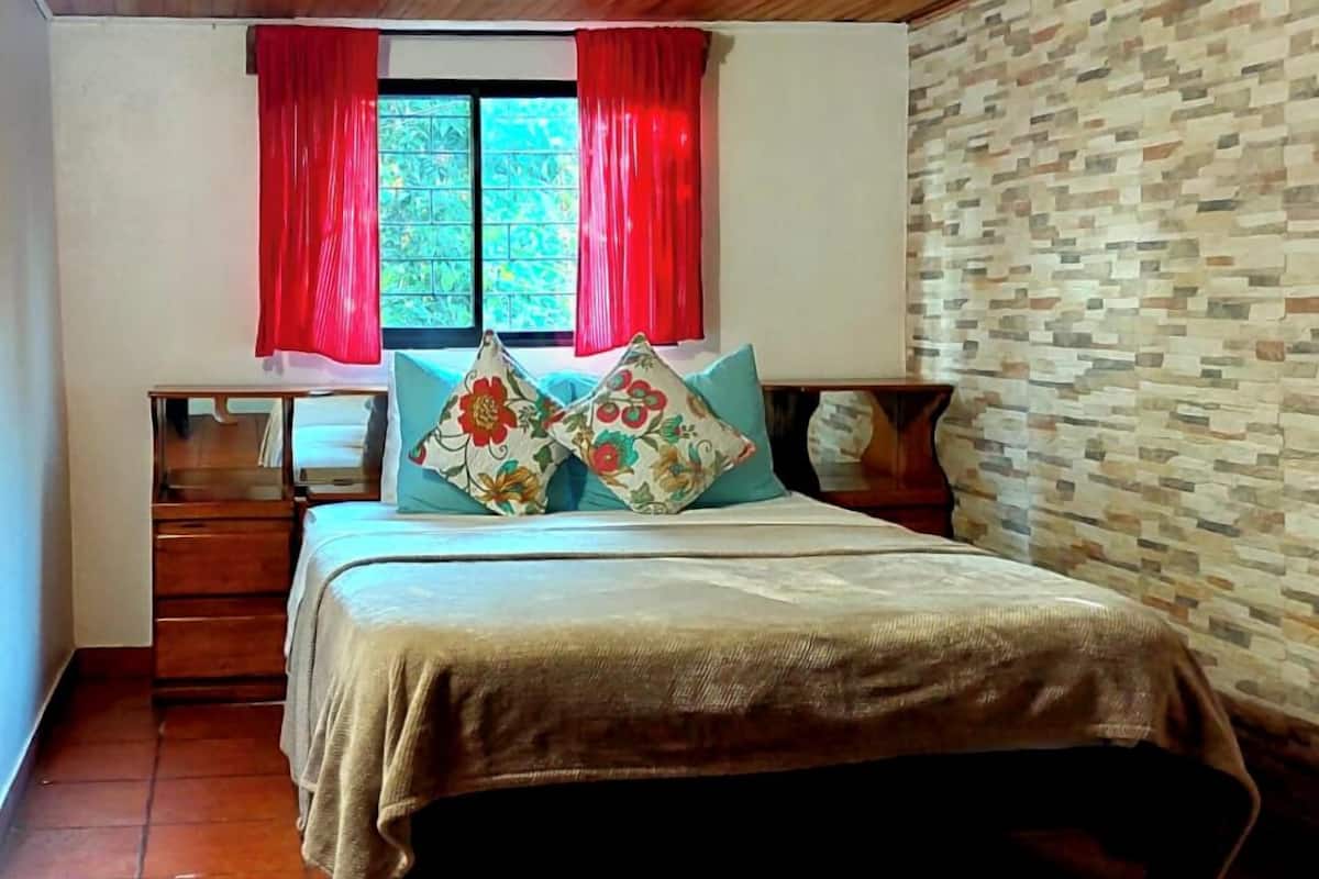 A bedroom with a wooden ceiling, a bed with floral pillows, a nightstand, a window with red curtains, and a tiled floor.