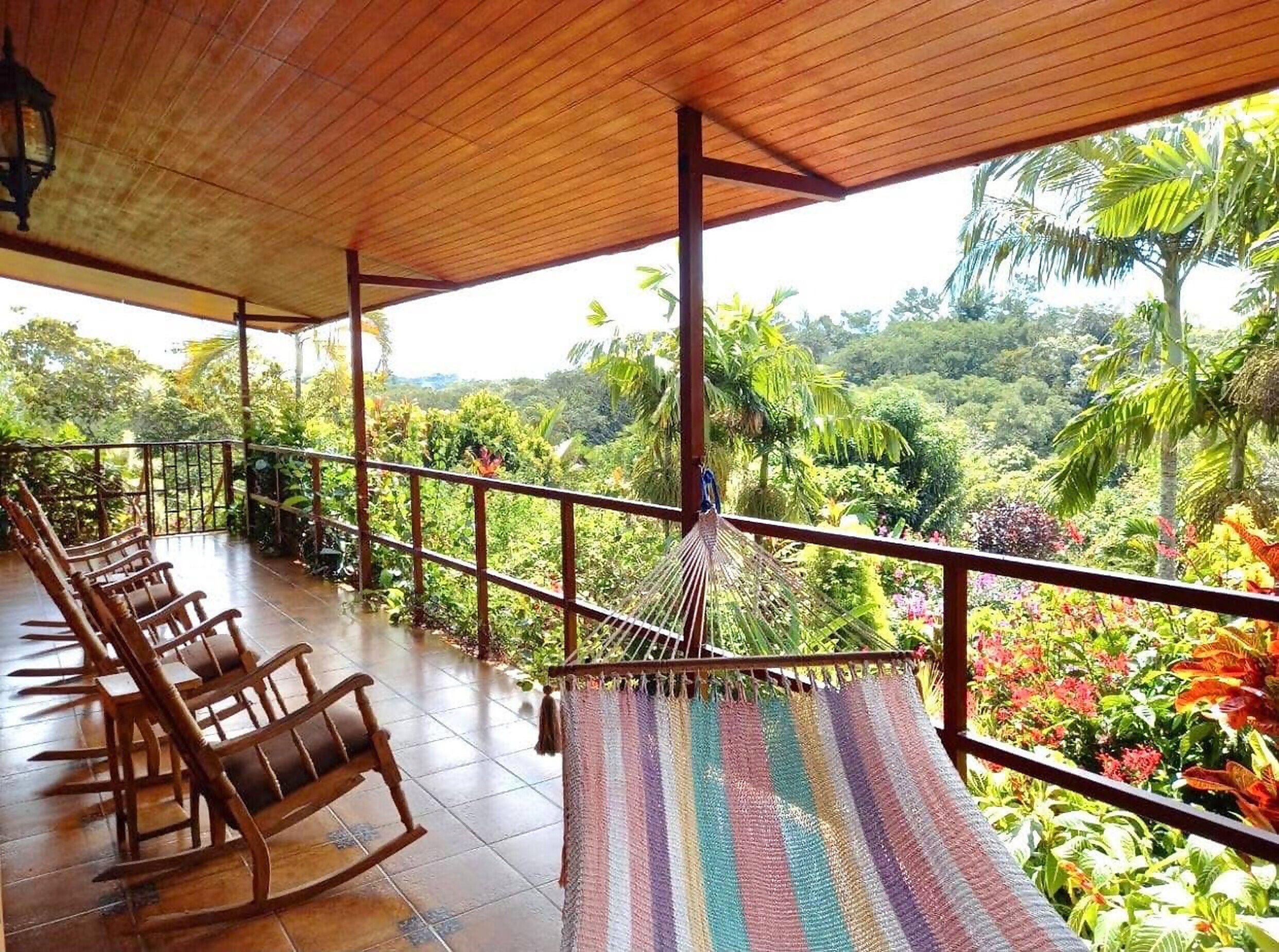 A covered outdoor seating area with wooden chairs and a hammock, overlooking a lush garden with various plants and trees.