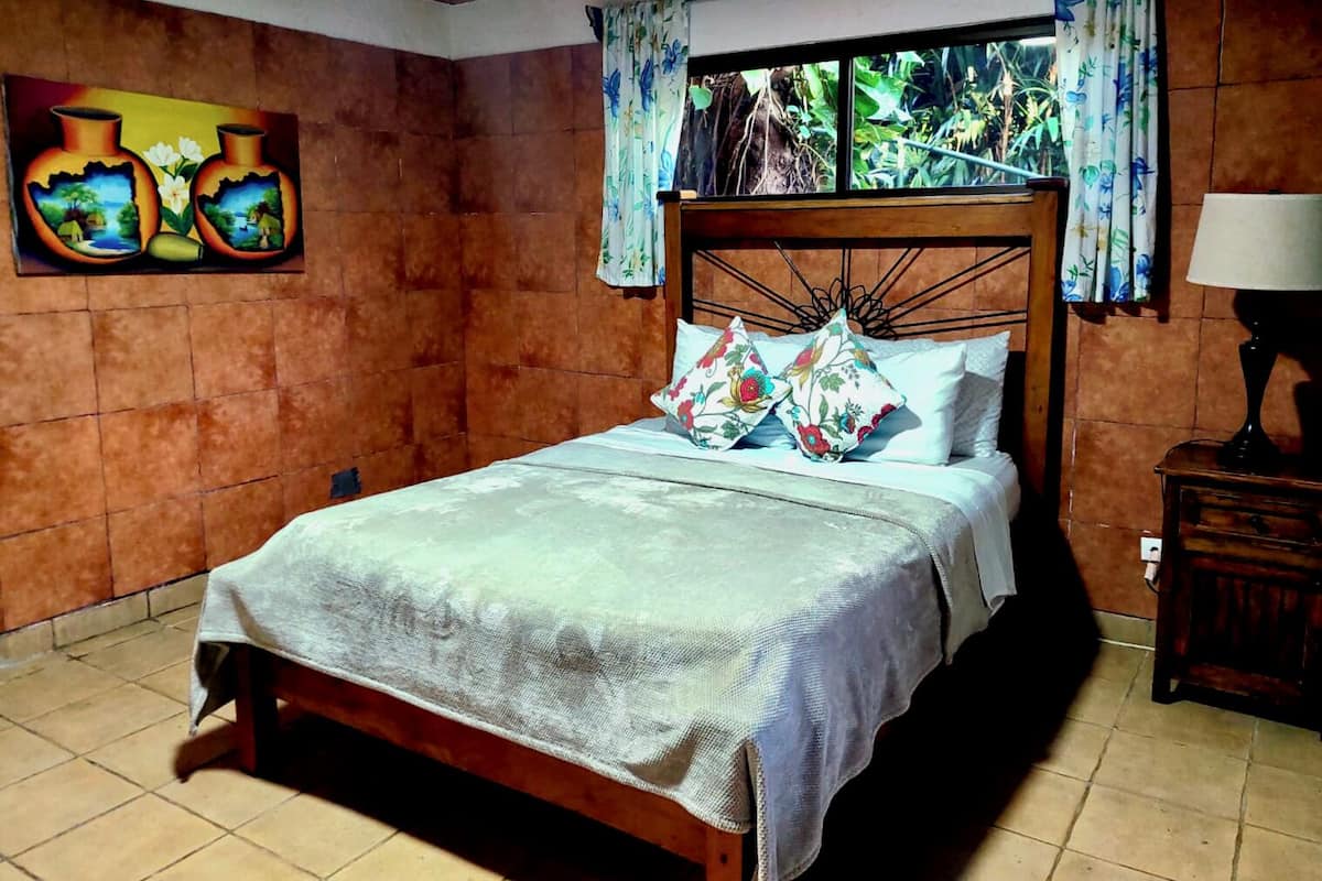 A bedroom with a wooden bed, floral pillows, a painting of vases, and a window with curtains.