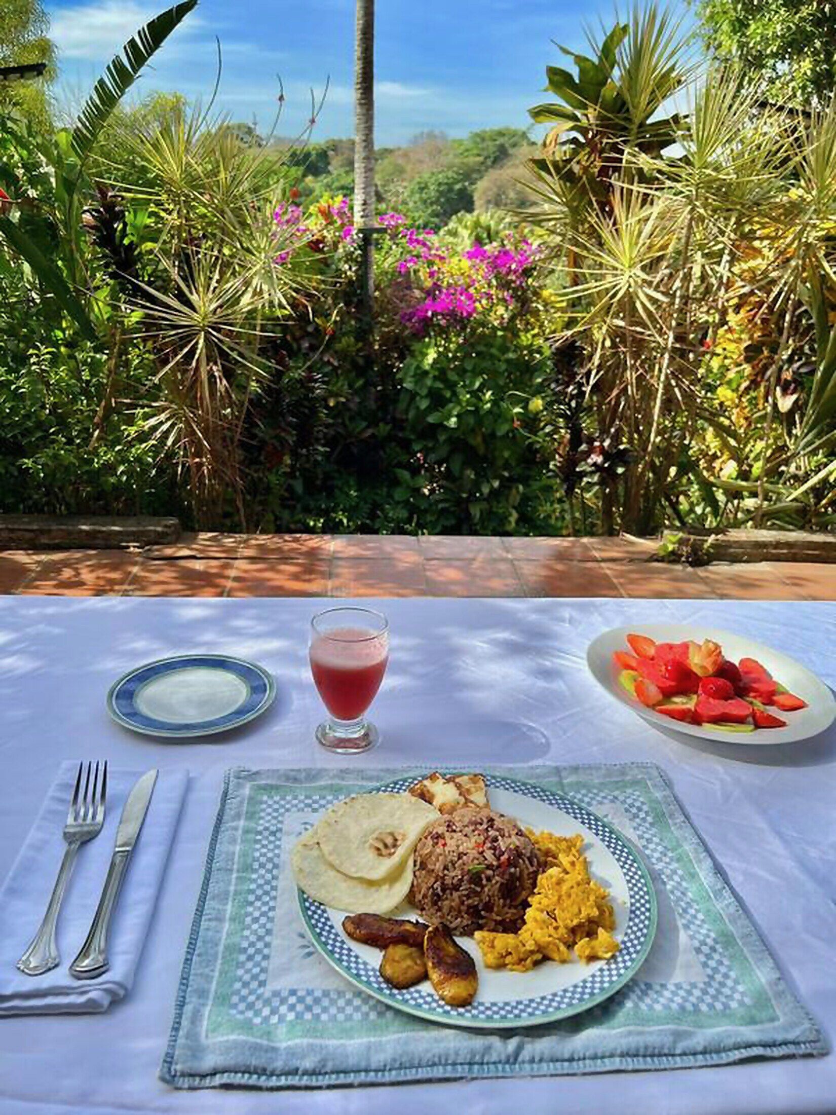 A table set outdoors with a meal consisting of rice, beans, and vegetables, a glass of red drink, and a plate of sliced fruit.