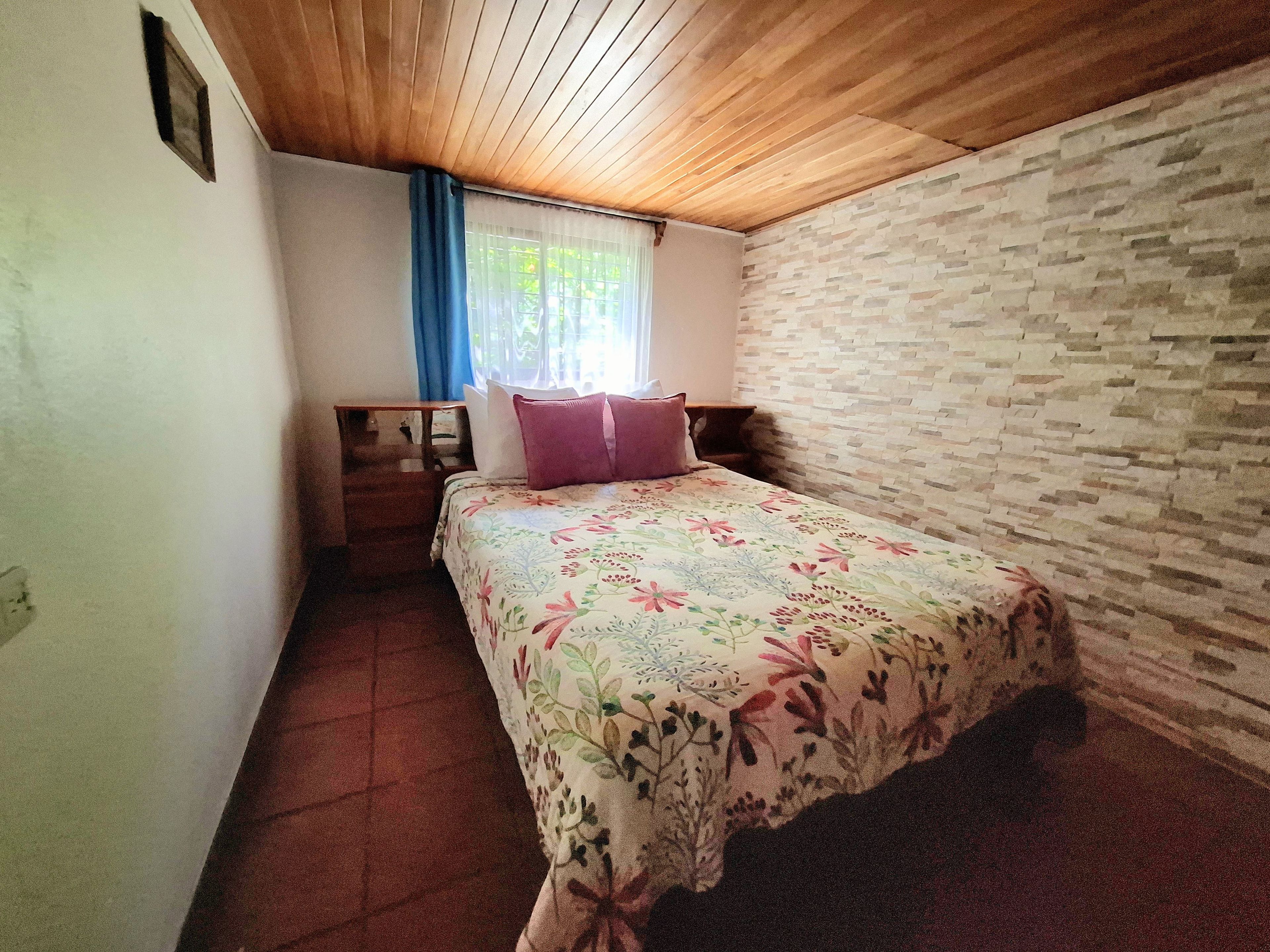 A bedroom with a wooden ceiling, a bed with floral bedding, a nightstand, and a window with blue curtains.