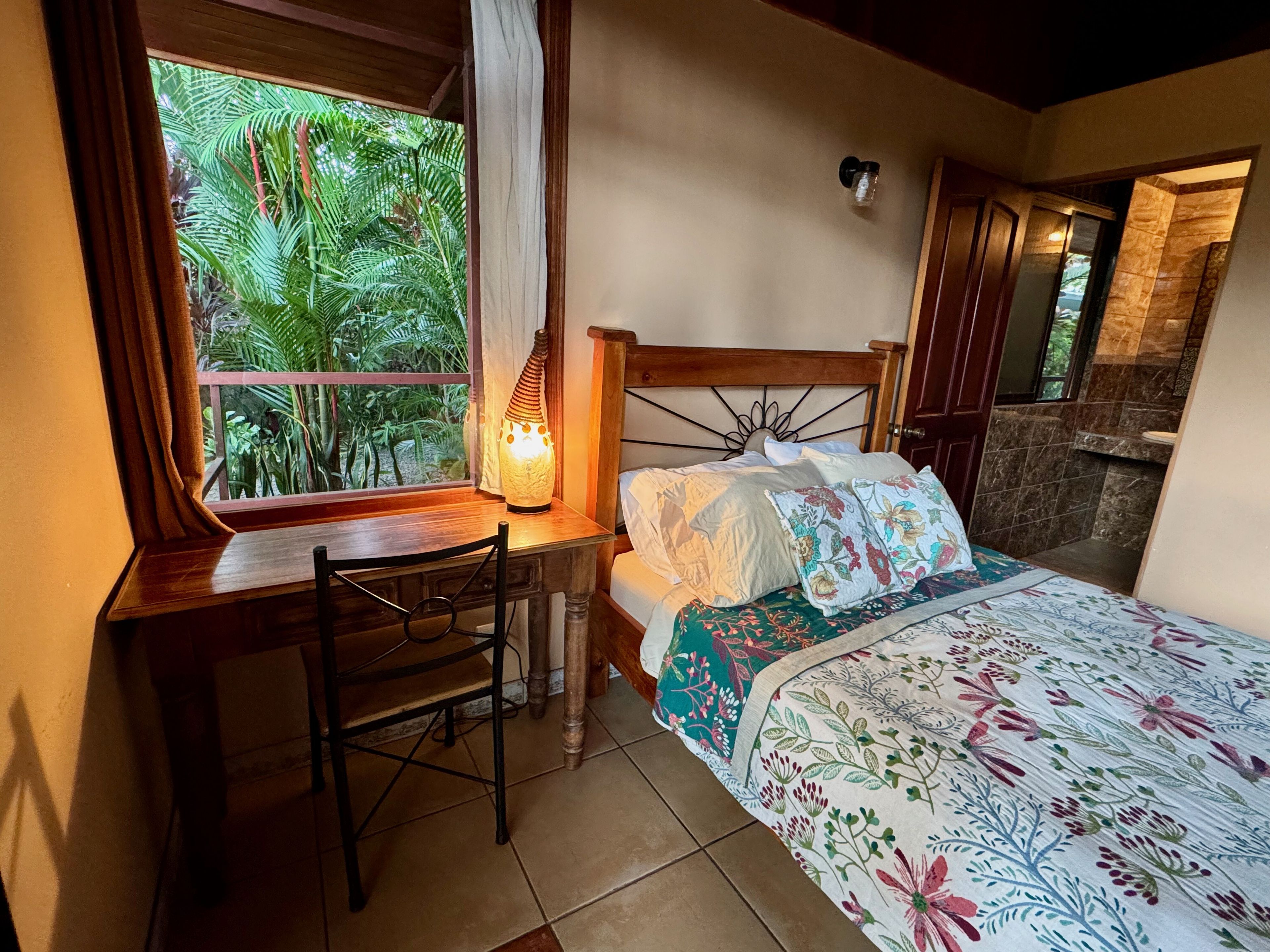 A bedroom with a wooden bed, a desk, and a chair. There is a window with curtains, a lamp, and a view of greenery outside.