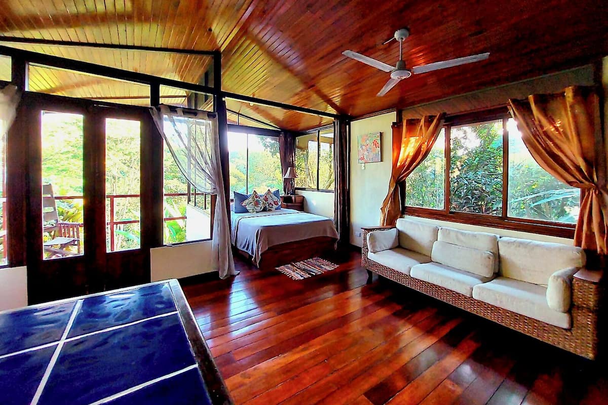 A room with a bed, a sofa, a ceiling fan, and large windows.