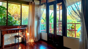 A room with wooden flooring, a wooden desk, and a view of greenery through large windows with curtains. - Eden Atenas (Mercedes)