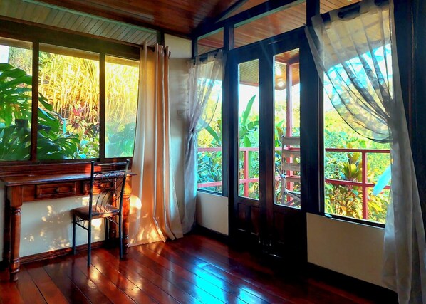 A room with wooden flooring, a wooden desk, and a view of greenery through large windows with curtains. - Eden Atenas (Mercedes)