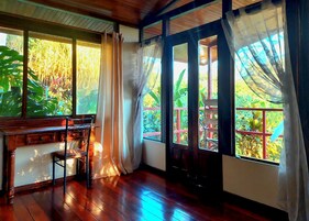 Quarto com piso de madeira, escrivaninha de madeira e vista para o verde através de amplas janelas com cortinas.