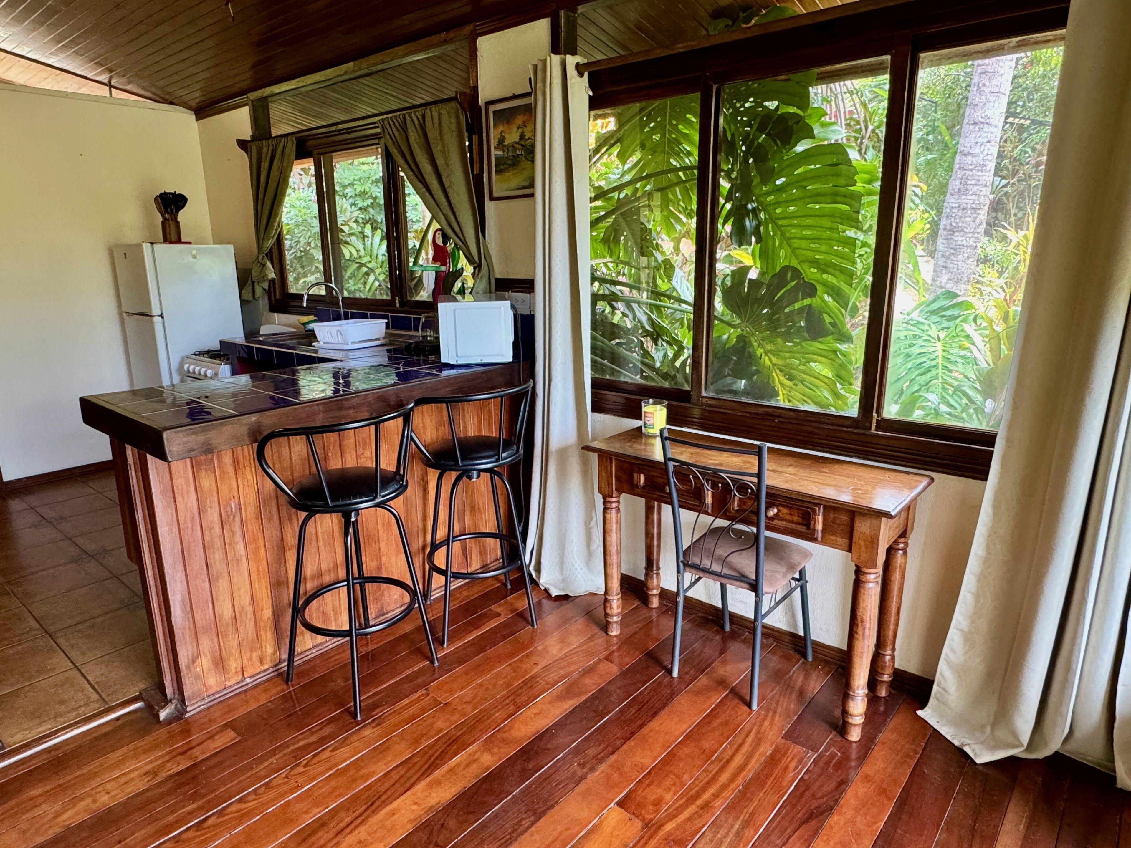 A kitchen with a wooden bar, stools, a refrigerator, and a window with a view of tropical foliage.