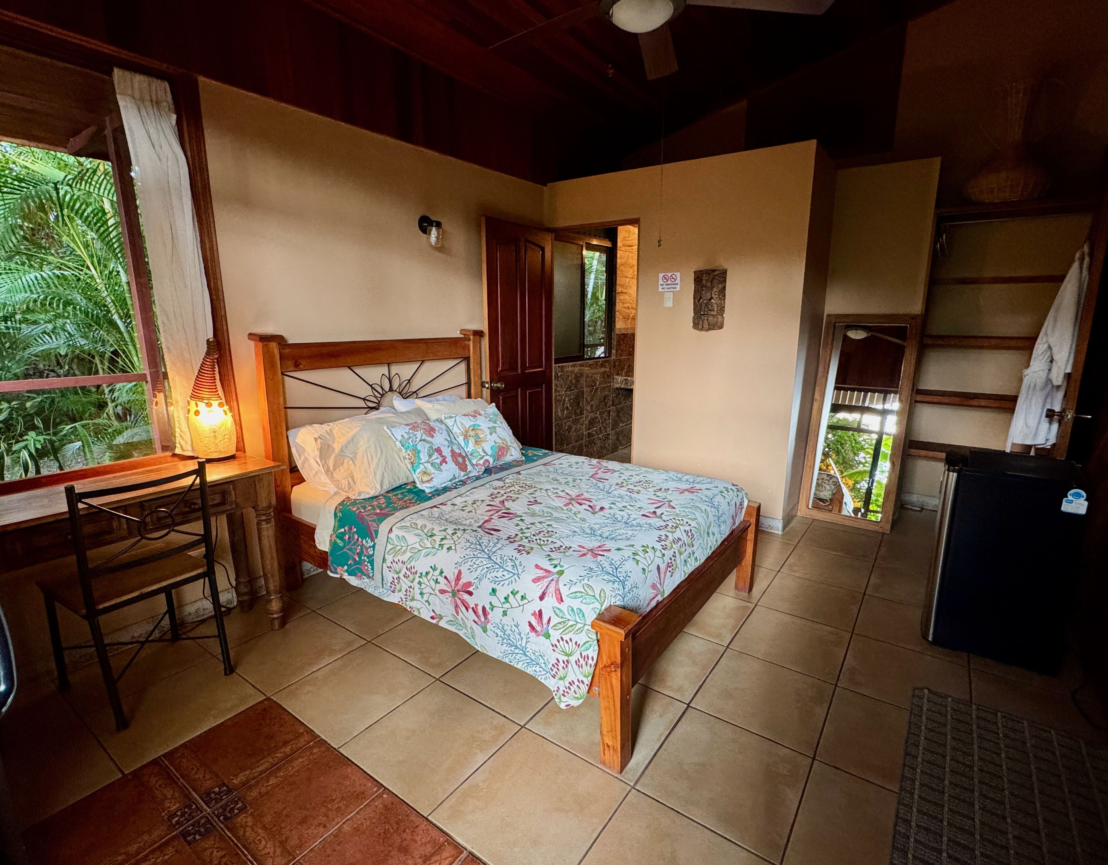 A bedroom with a wooden bed, a desk with a lamp, and a view of greenery outside.