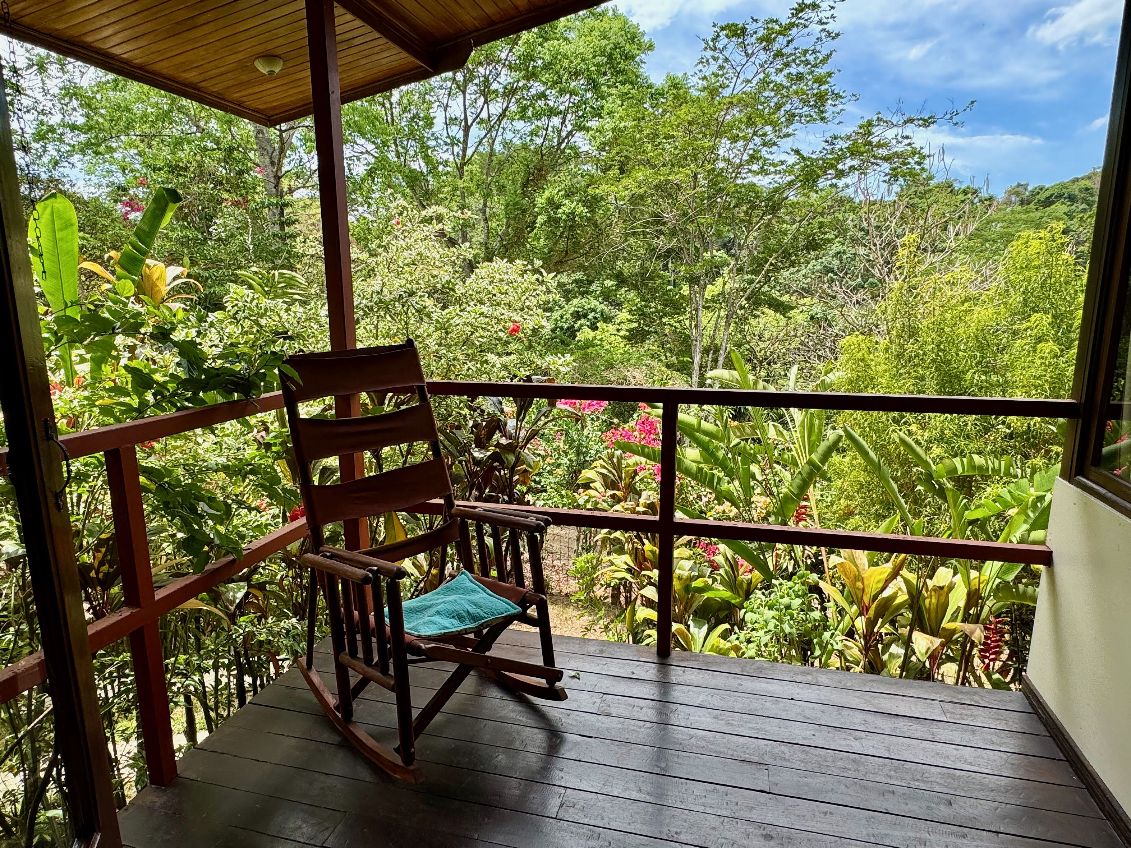 Una terraza de madera con una mecedora, rodeada de abundante follaje y árboles.