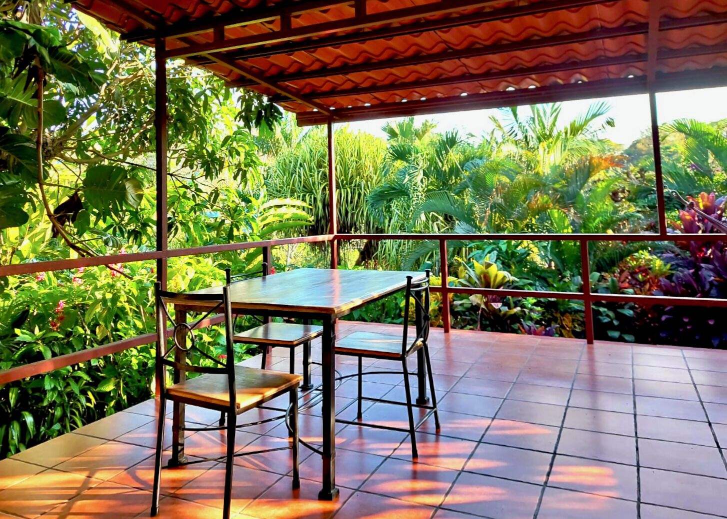 Uma área de estar coberta ao ar livre, com mesa e cadeiras, rodeada por vegetação tropical exuberante.