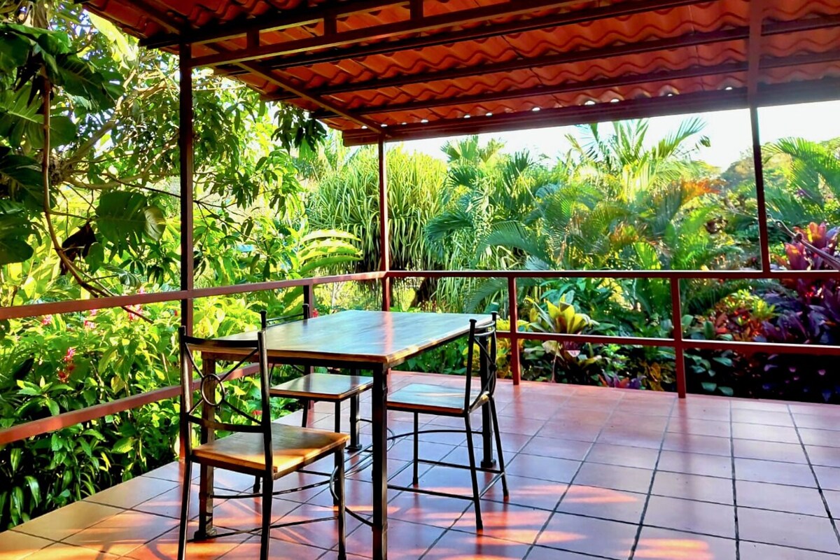A covered outdoor seating area with a table and chairs, surrounded by lush tropical vegetation.