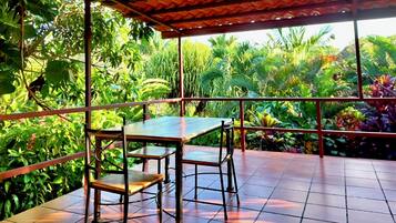 Uma área de estar coberta ao ar livre, com mesa e cadeiras, rodeada por vegetação tropical exuberante.