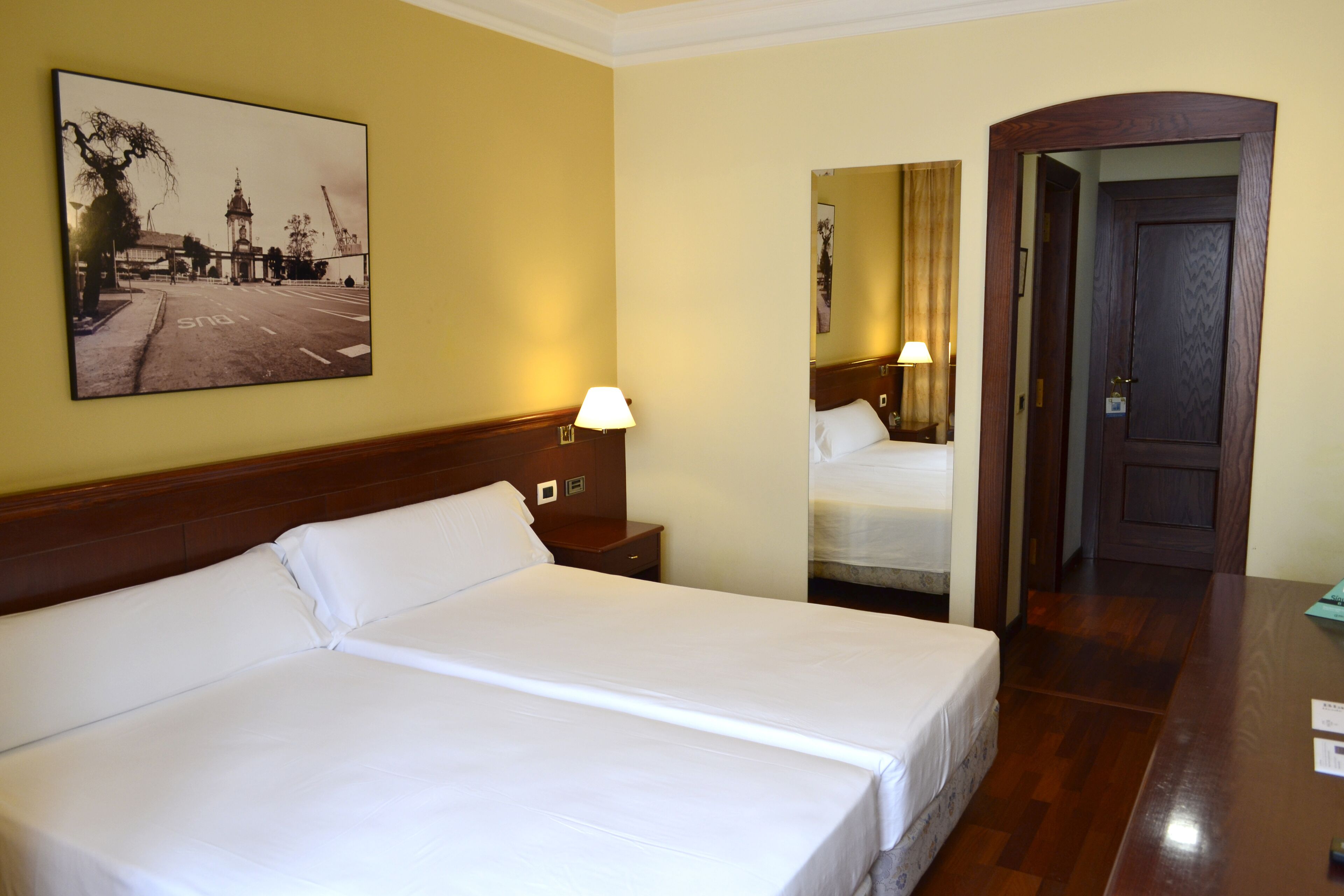 Habitación de hotel con una cama grande, una mesita de noche con lámpara, un espejo y una puerta que da a otra habitación.