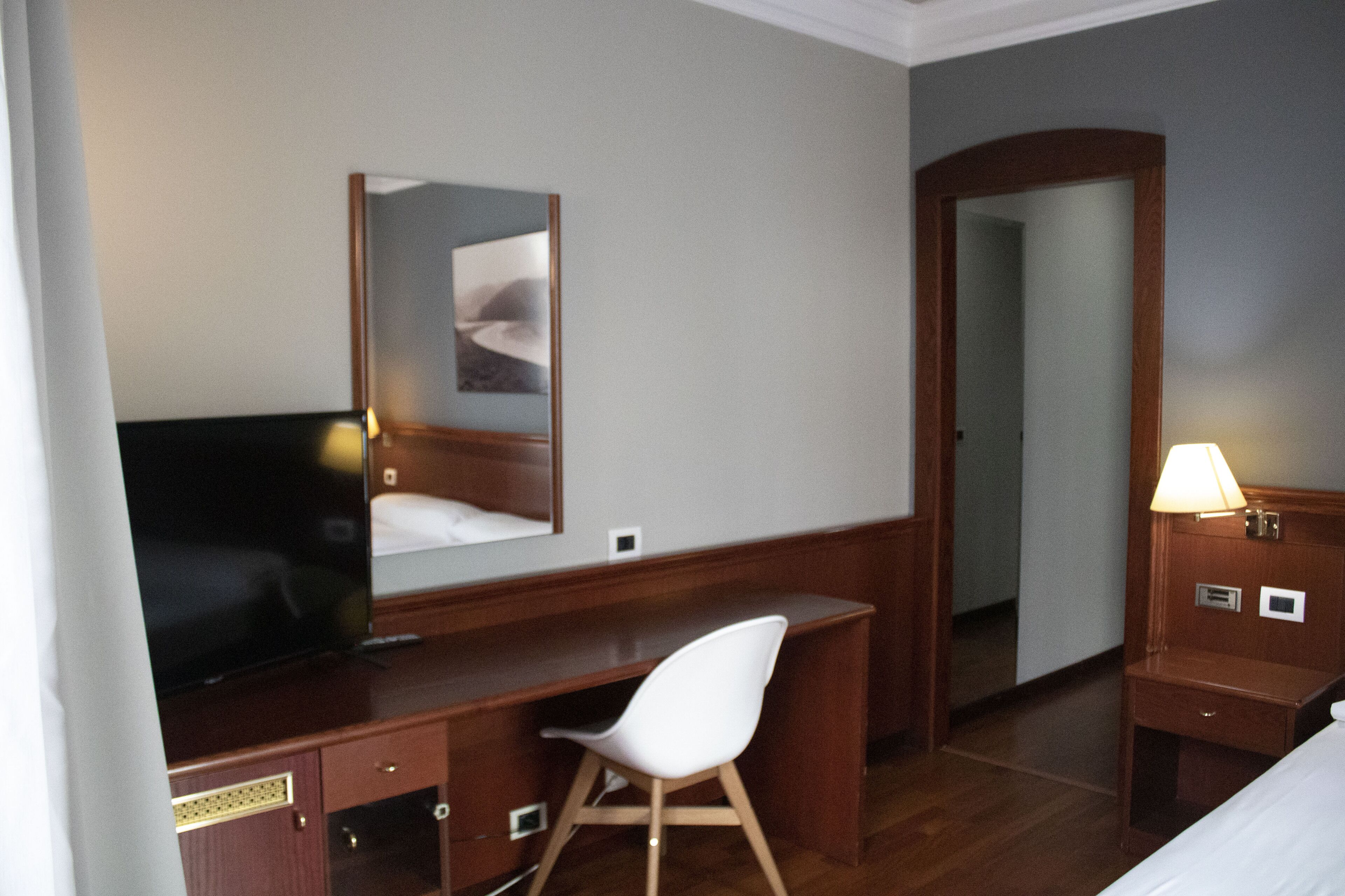 Une chambre d’hôtel avec un bureau en bois, une chaise blanche, une télévision, un miroir, une table de chevet avec une lampe et une porte donnant sur une autre pièce.