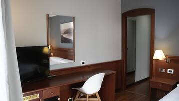 Une chambre d’hôtel avec un bureau en bois, une chaise blanche, une télévision, un miroir, une table de chevet avec une lampe et une porte donnant sur une autre pièce.