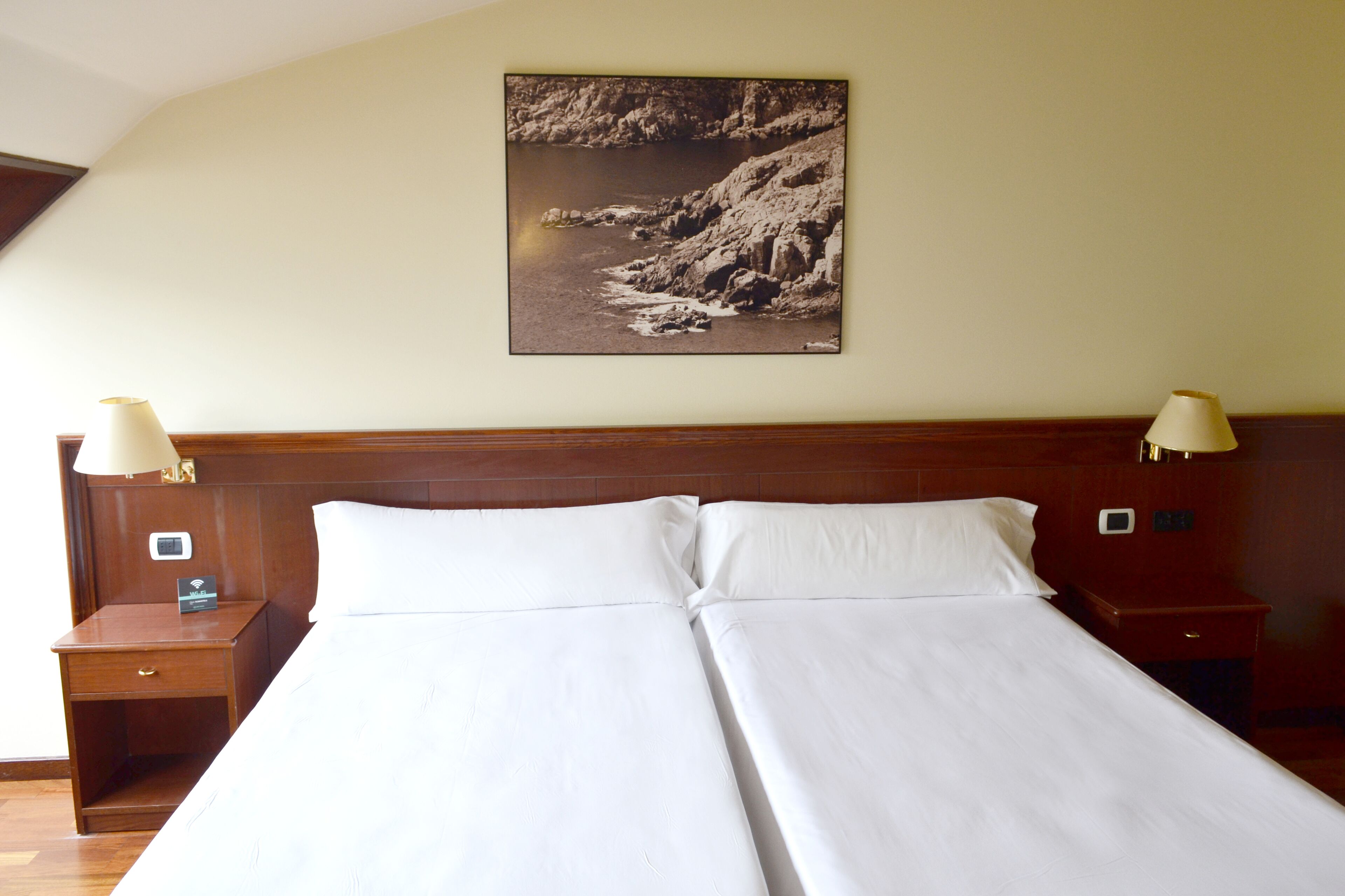 Un lit double avec du linge de lit blanc, deux tables de chevet avec des lampes, une petite table de nuit avec une horloge et, au mur, un tableau représentant un paysage côtier.