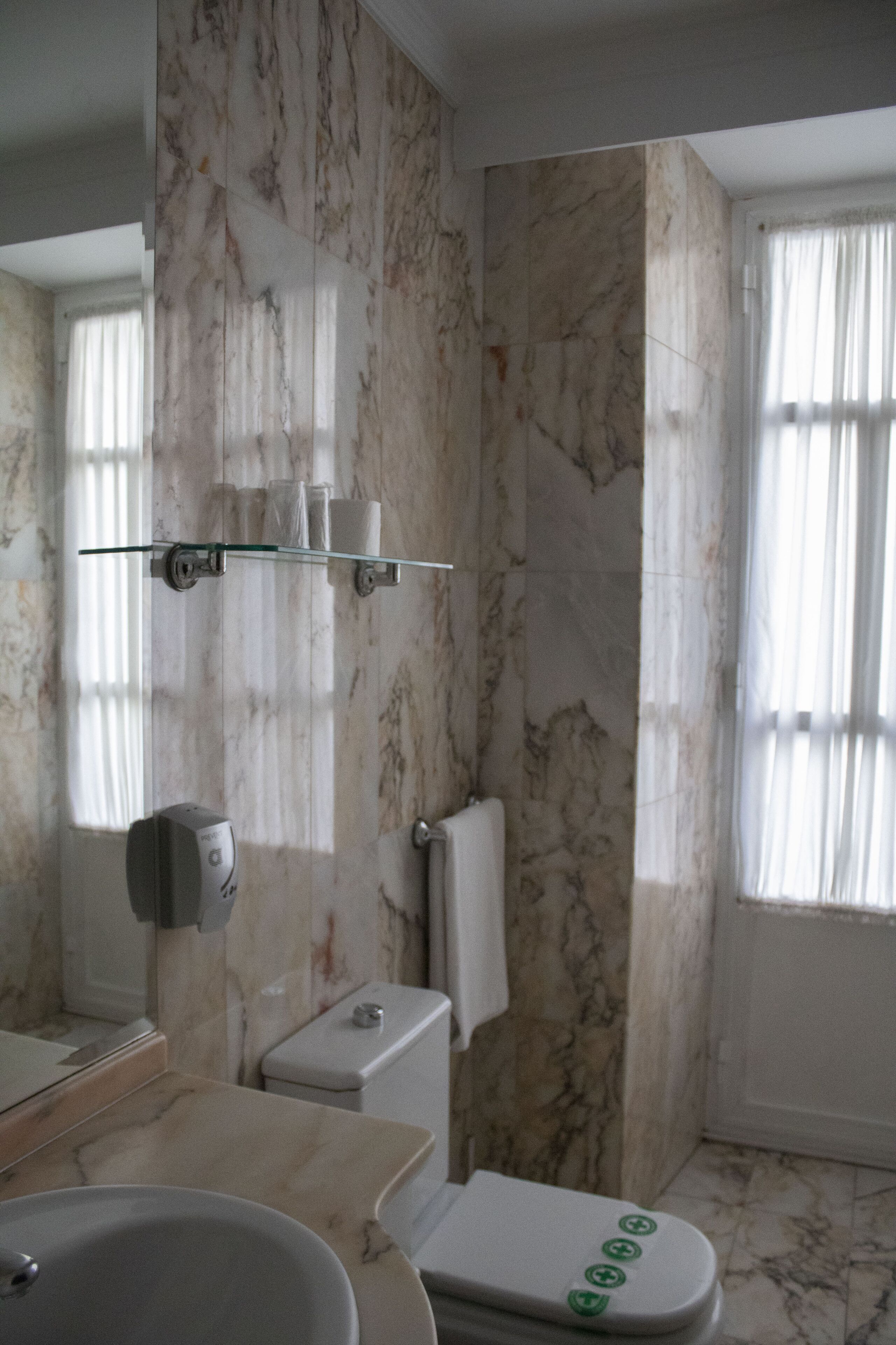 A bathroom with a marble wall, a glass shelf with toiletries, a toilet, and a sink.