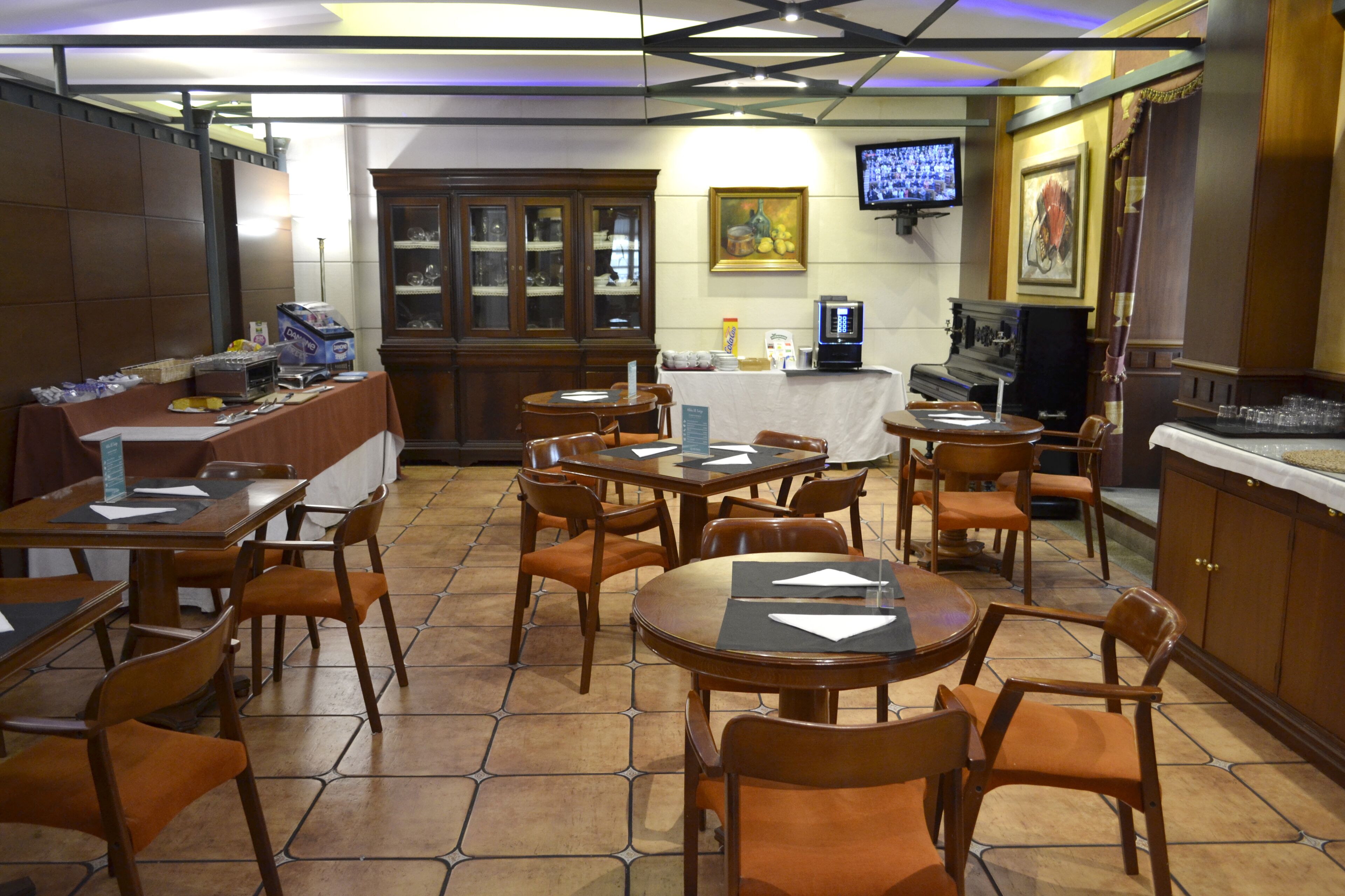 A restaurant with multiple tables and chairs, a wooden cabinet, a television mounted on the wall, and a tiled floor.