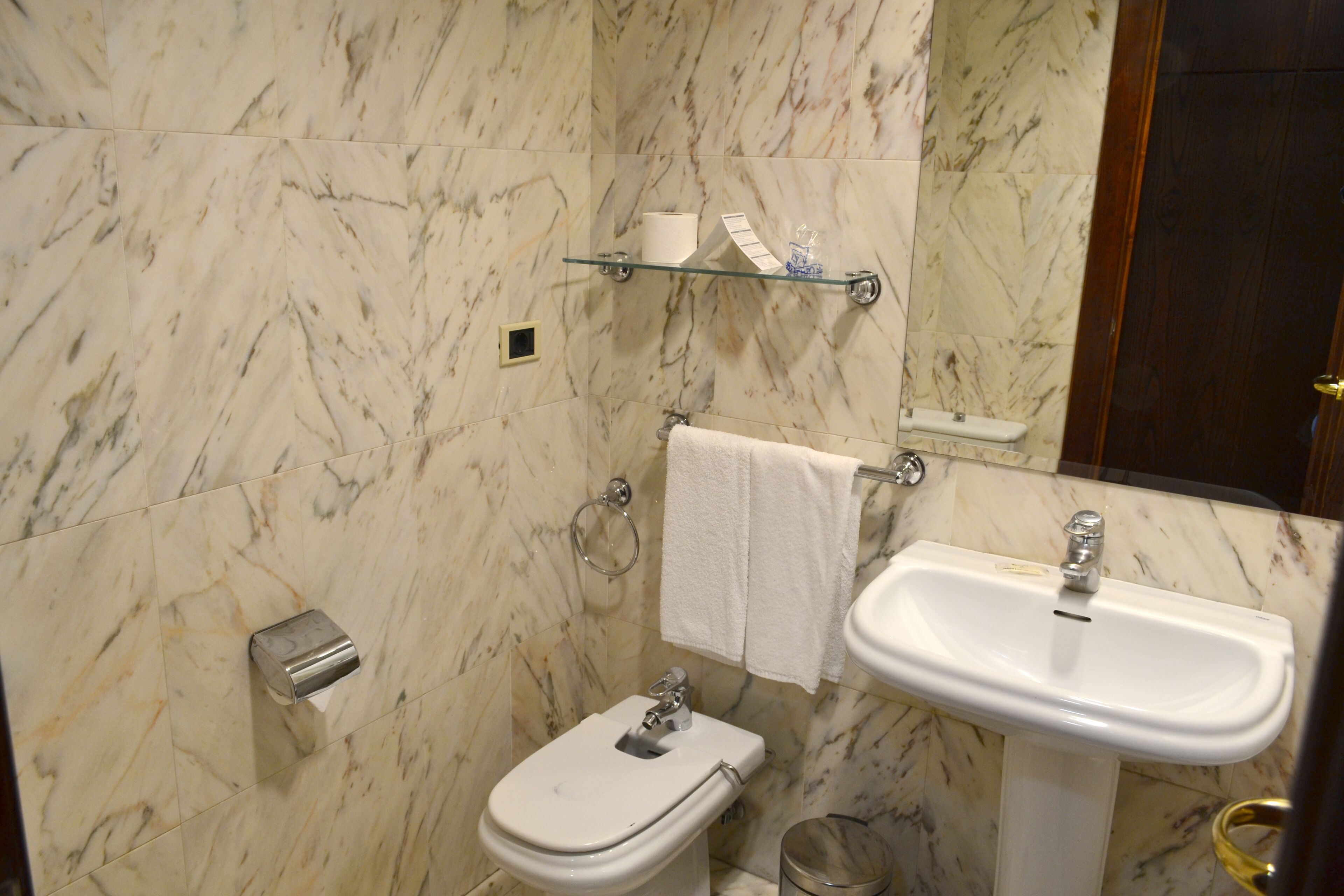 Une salle de bain avec un mur en marbre, des toilettes, un lavabo, une étagère en verre où sont rangés les articles de toilette et un porte-serviettes.