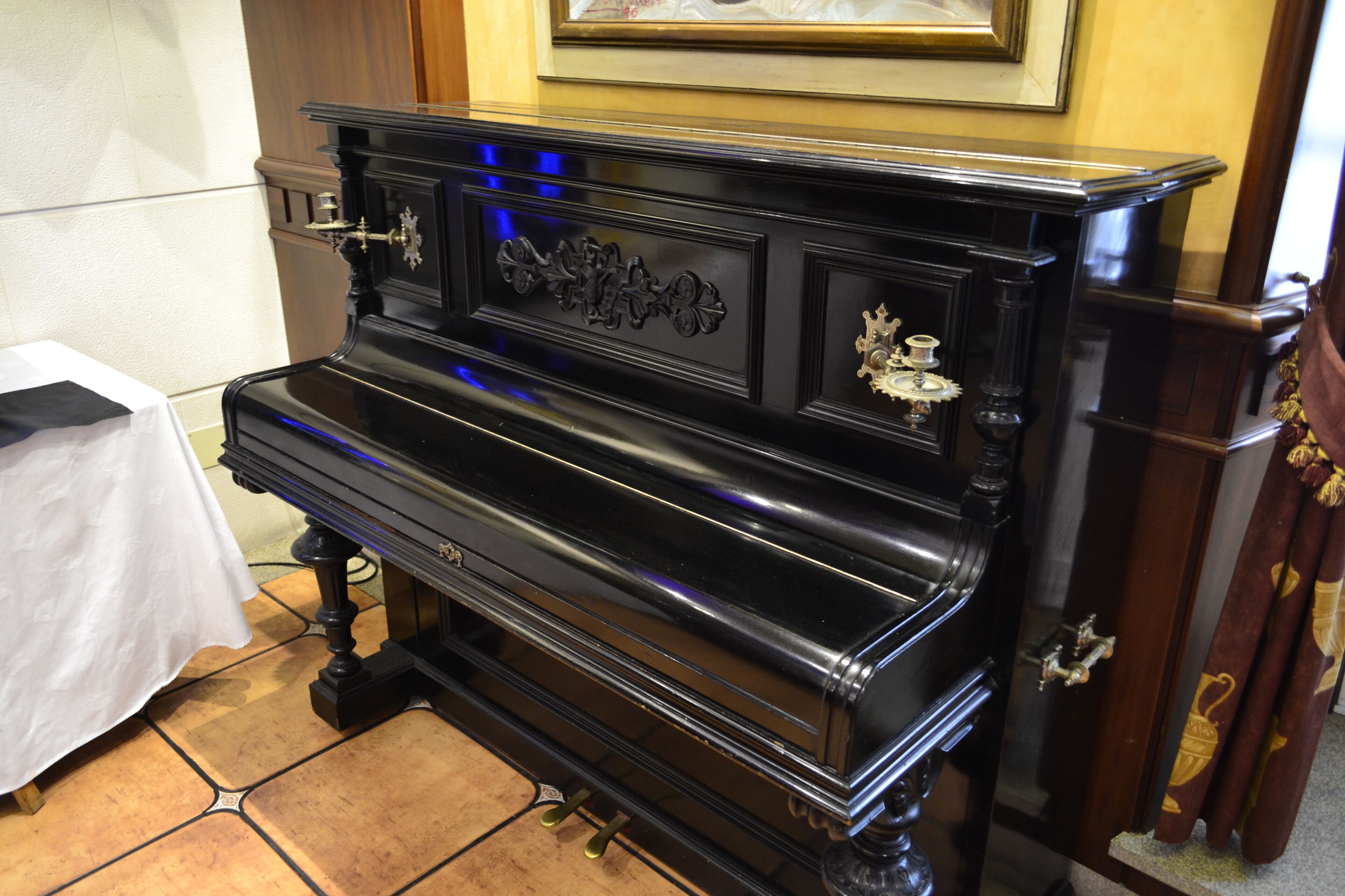 A black upright piano with ornate carvings and brass fittings, positioned in a room with tiled flooring and a white tablecloth-covered table to the left.